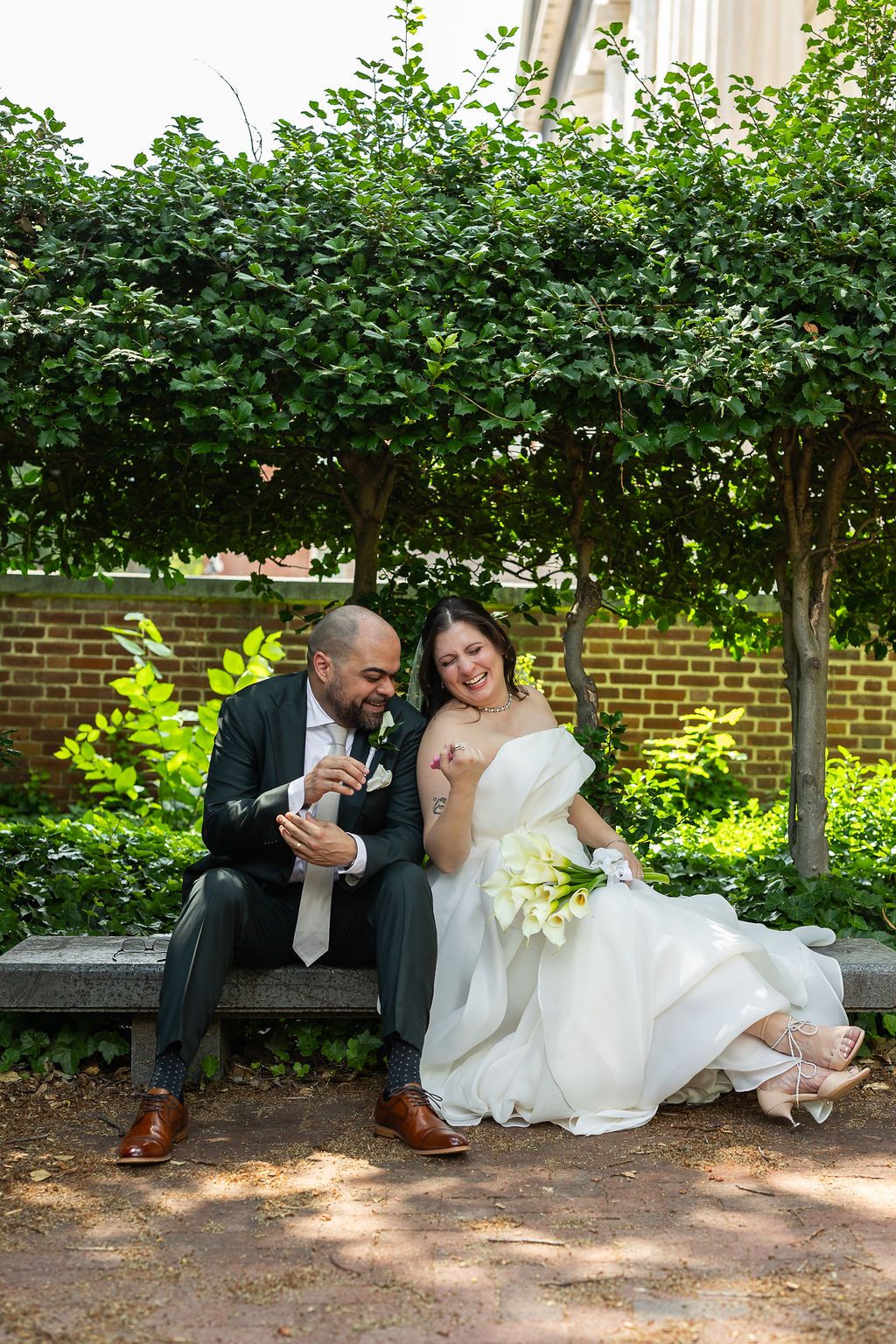 Bride and groom sitting on a garden bench laughing together, bride holding white calla lily bouquet in wedding dress.