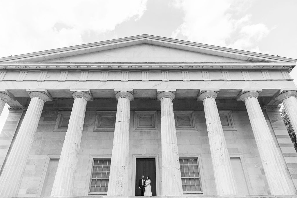 Bride and groom posing beneath towering classical columns of a historic building in black and white.