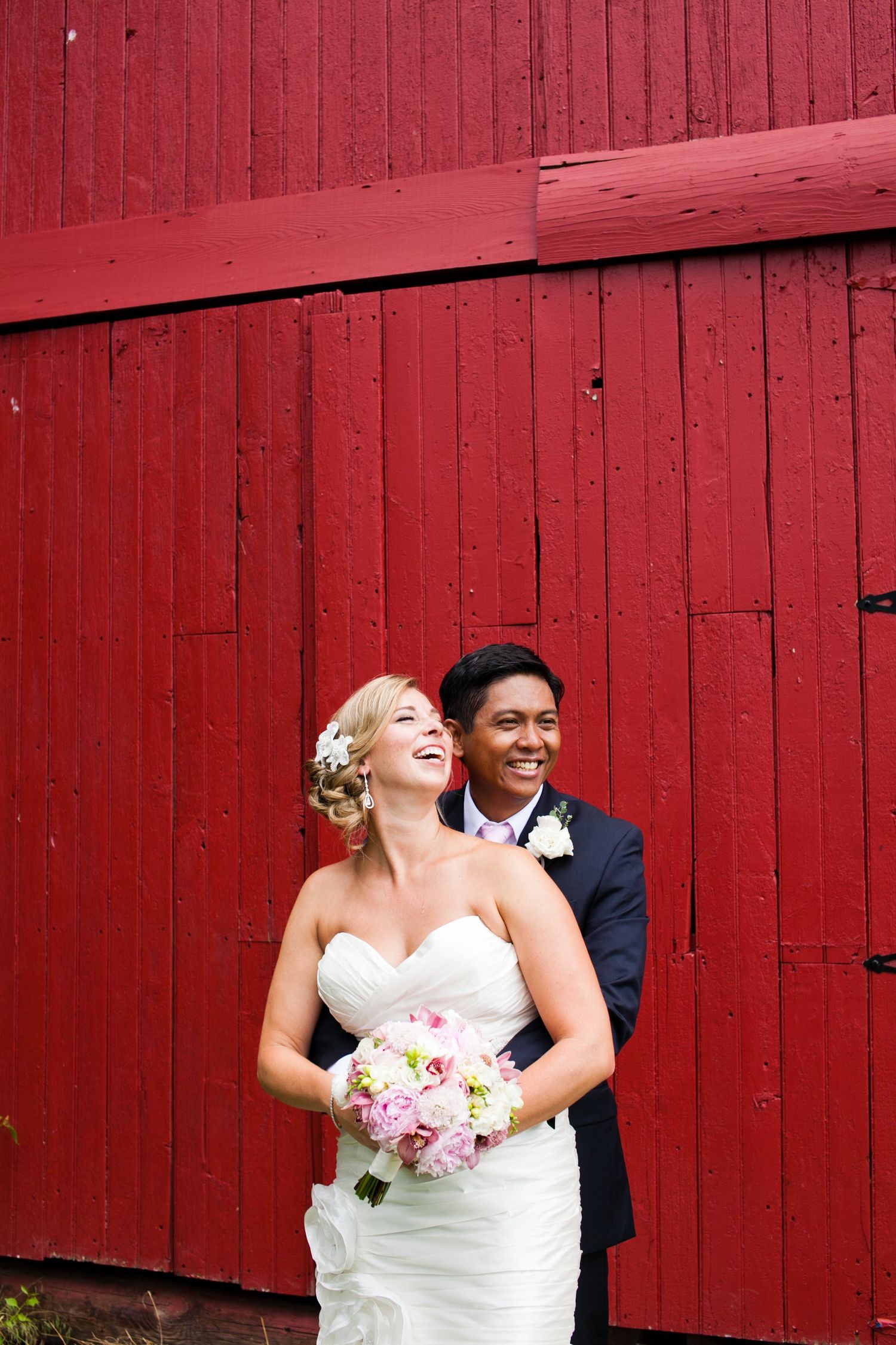 Happy bride and groom laughing together in front of red barn wall.