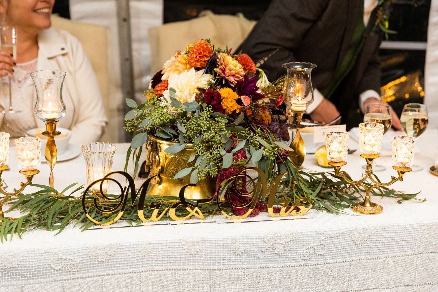 Lush autumn wedding centerpiece with orange, burgundy, and white blooms on gold candlelit table.