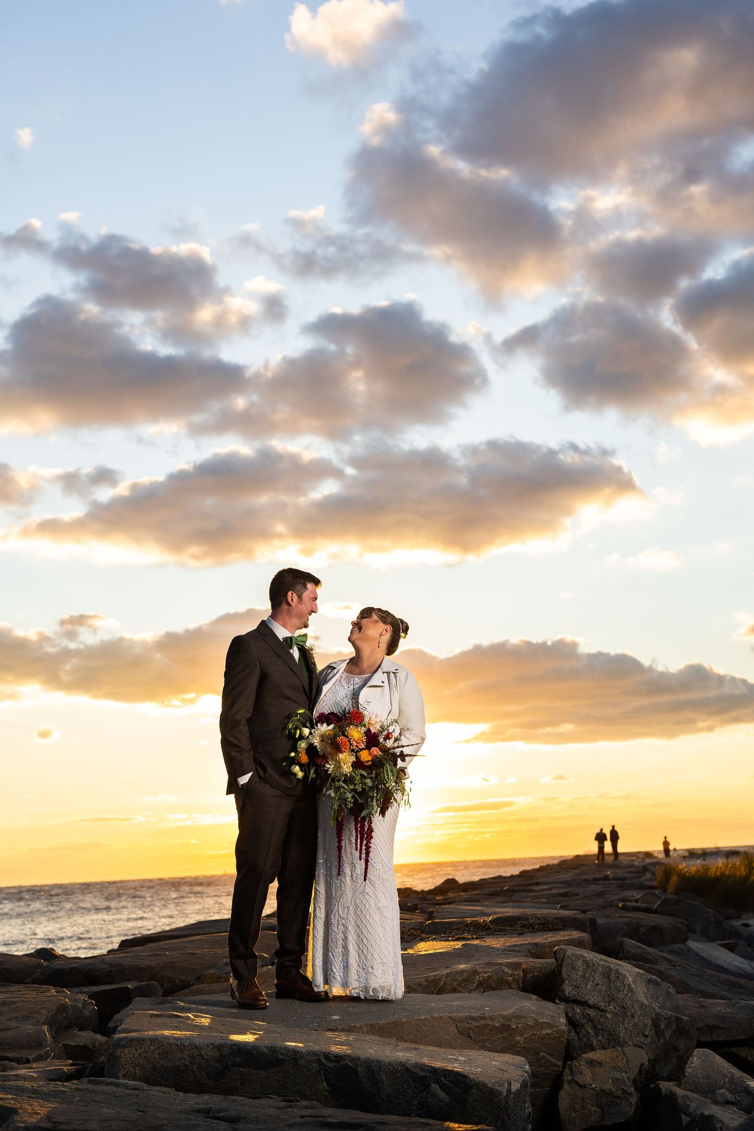 Bride and groom share romantic gaze on rocky coast beneath vibrant golden sunset clouds.