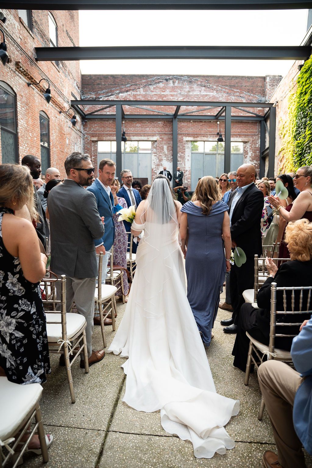 Bride in white gown with veil walks down aisle at industrial outdoor wedding ceremony with brick walls.
