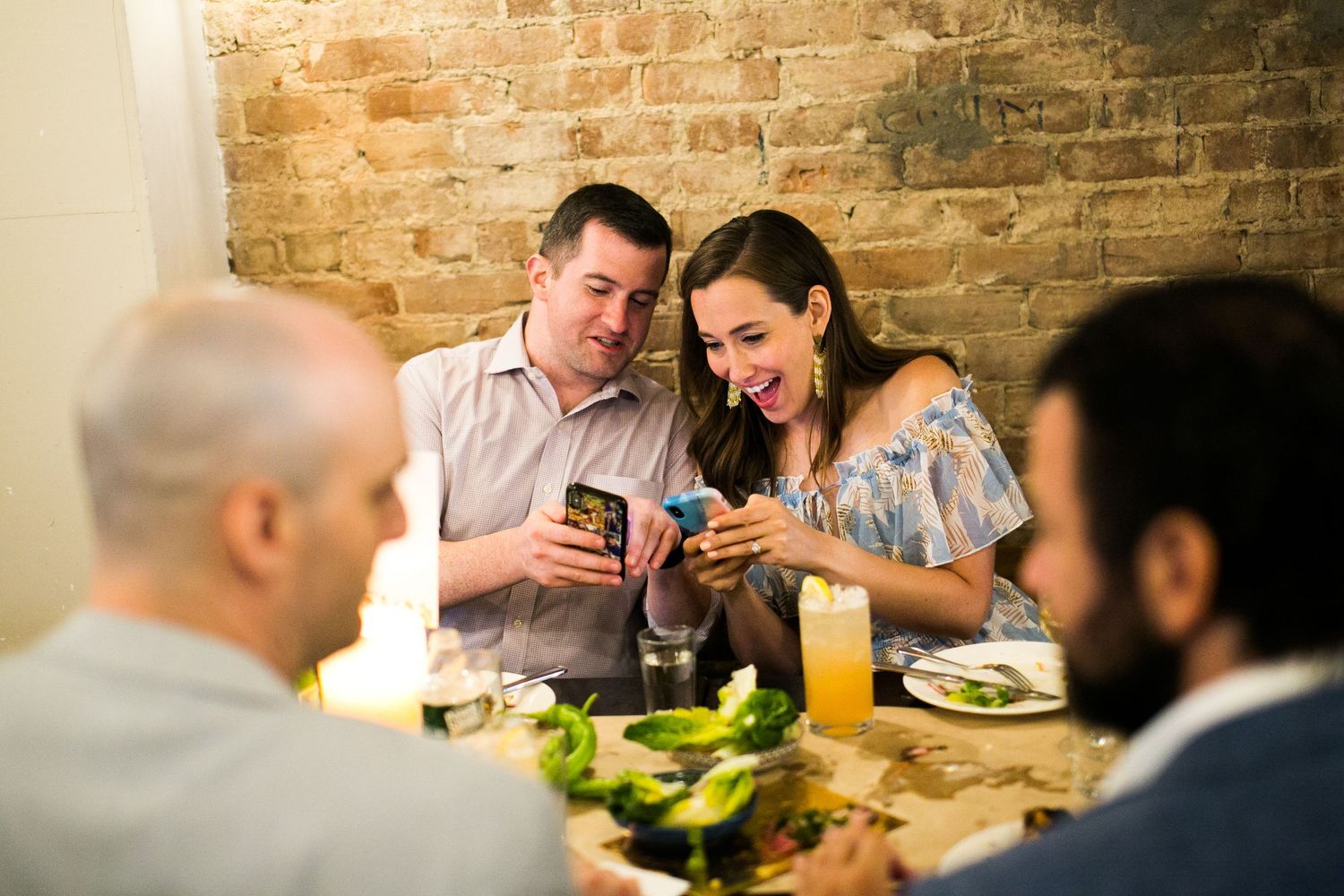 Couple smiles while looking at phone together during candlelit dinner at rustic brick wall restaurant.