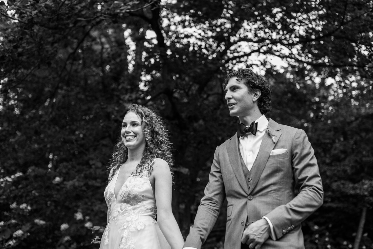 Black and white portrait of smiling bride and groom holding hands outdoors among trees.