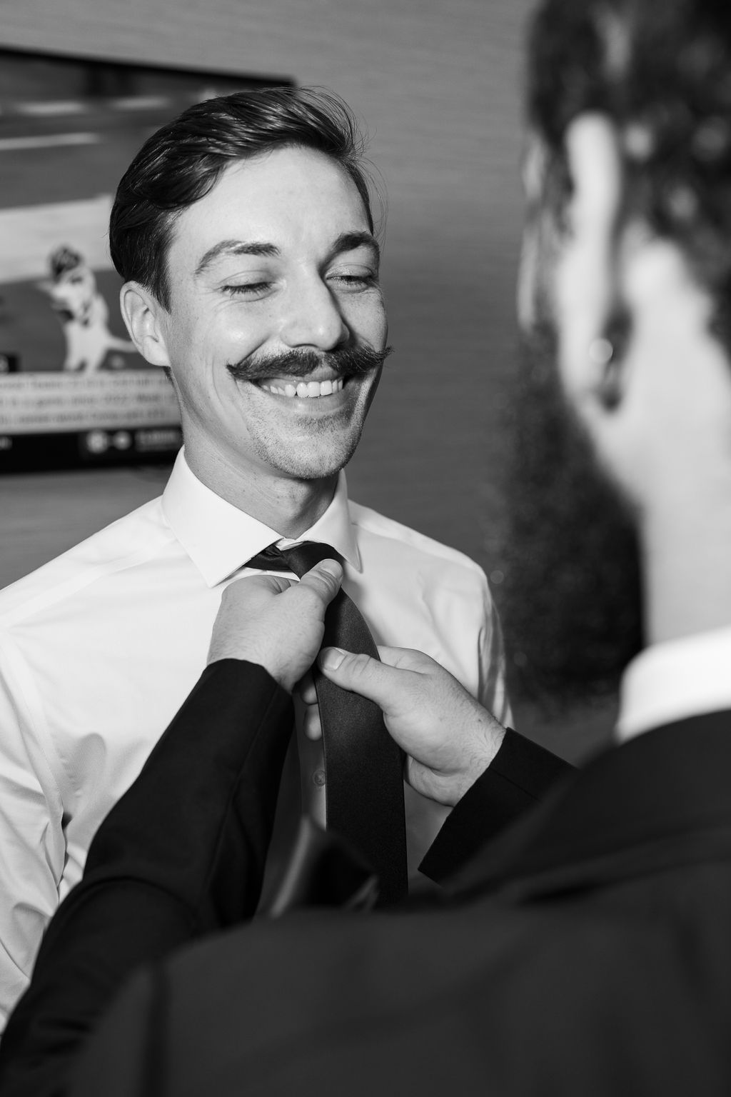 Black and white photo of a smiling groom with mustache having his tie adjusted by a groomsman.