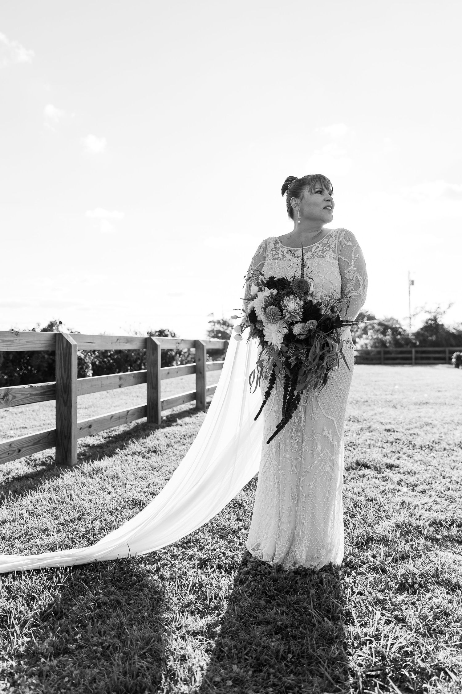 Bride in lace wedding gown with long veil standing by rustic fence, black and white outdoor portrait.