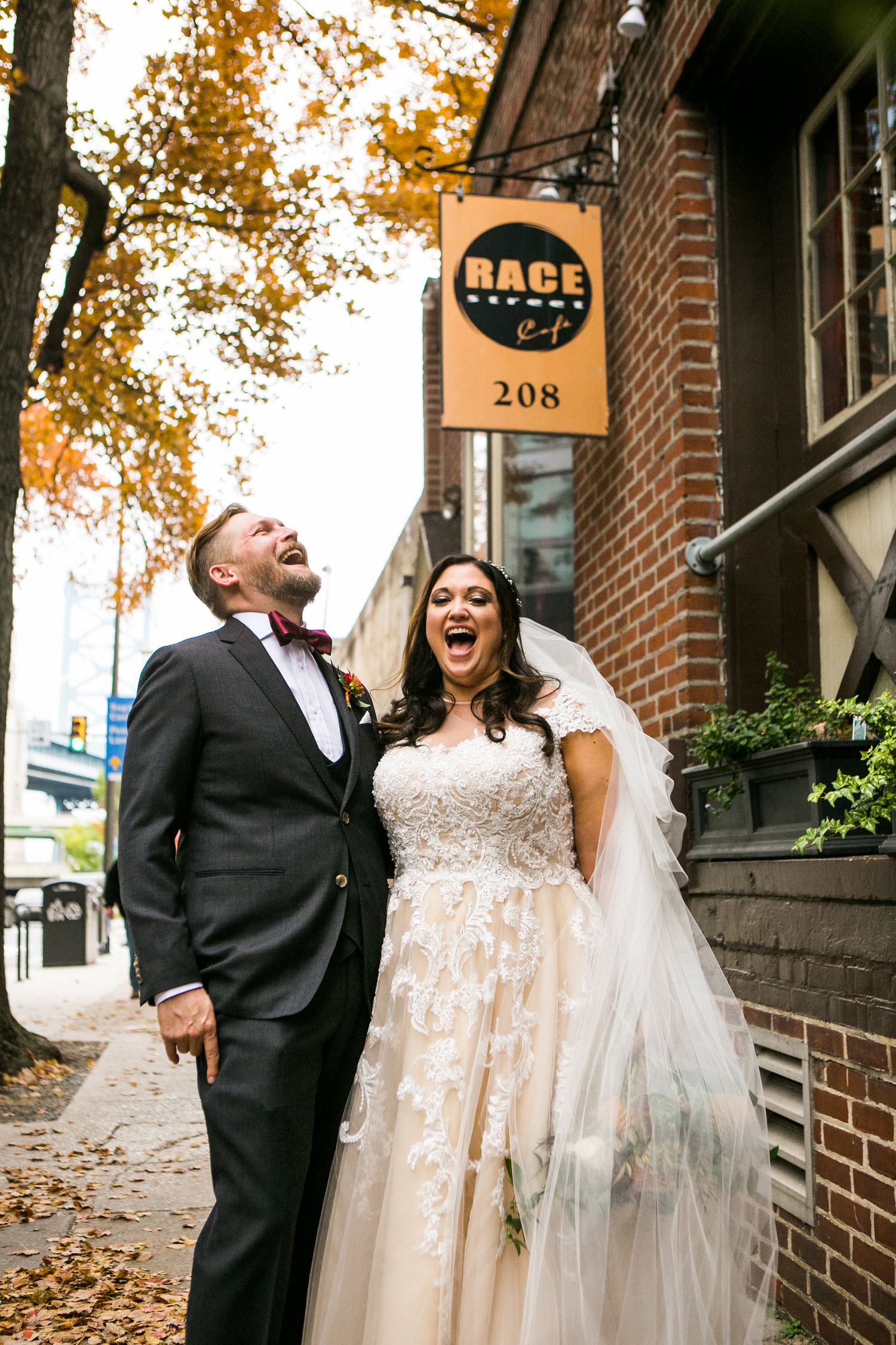 Laughing bride and groom pose joyfully outside Race Street Cafe sign with fall foliage backdrop.