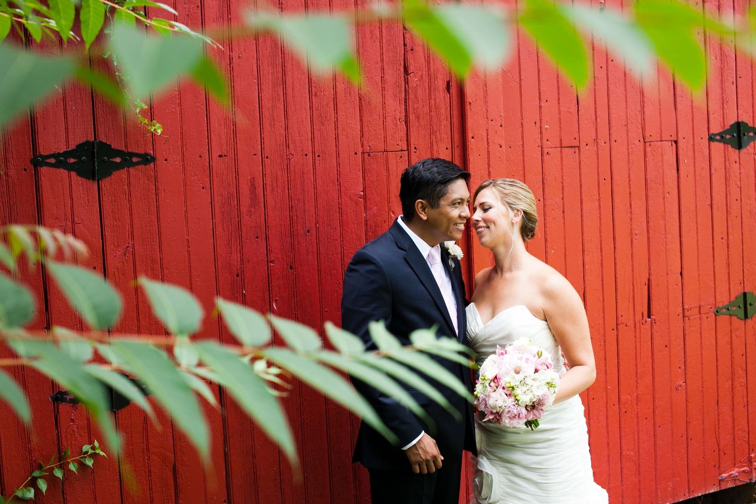 Interracial couple sharing romantic moment by red barn wall with greenery.