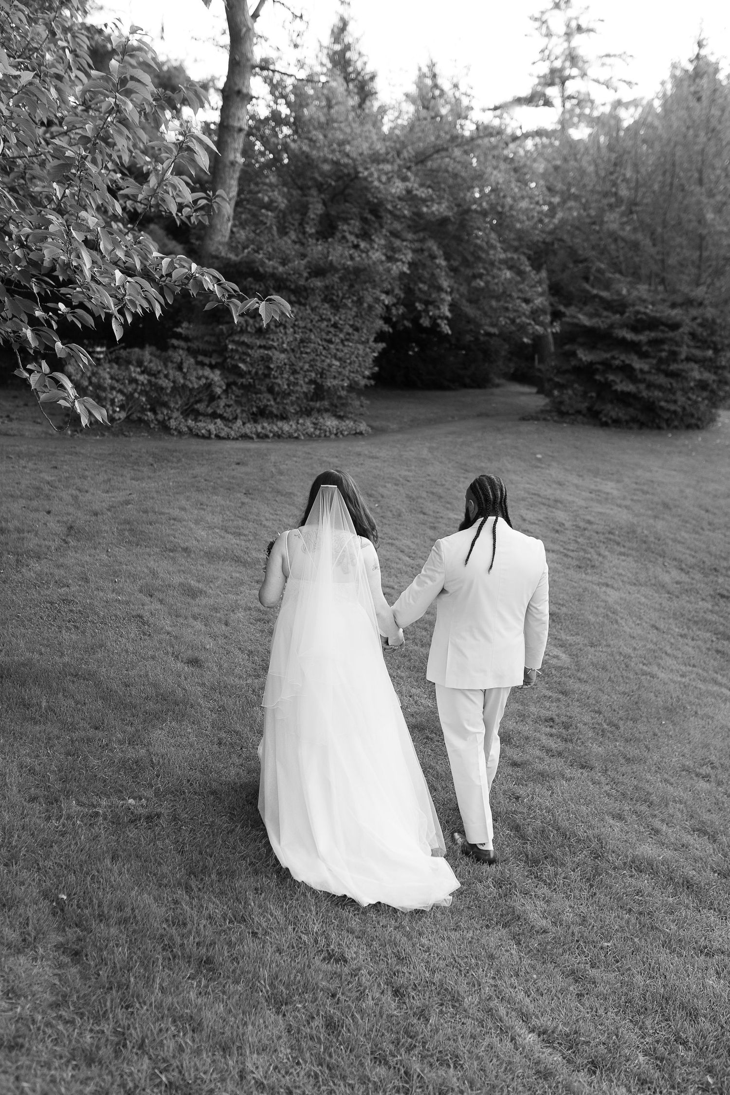 Black and white rear view of bride and groom walking hand in hand across a lush green lawn.
