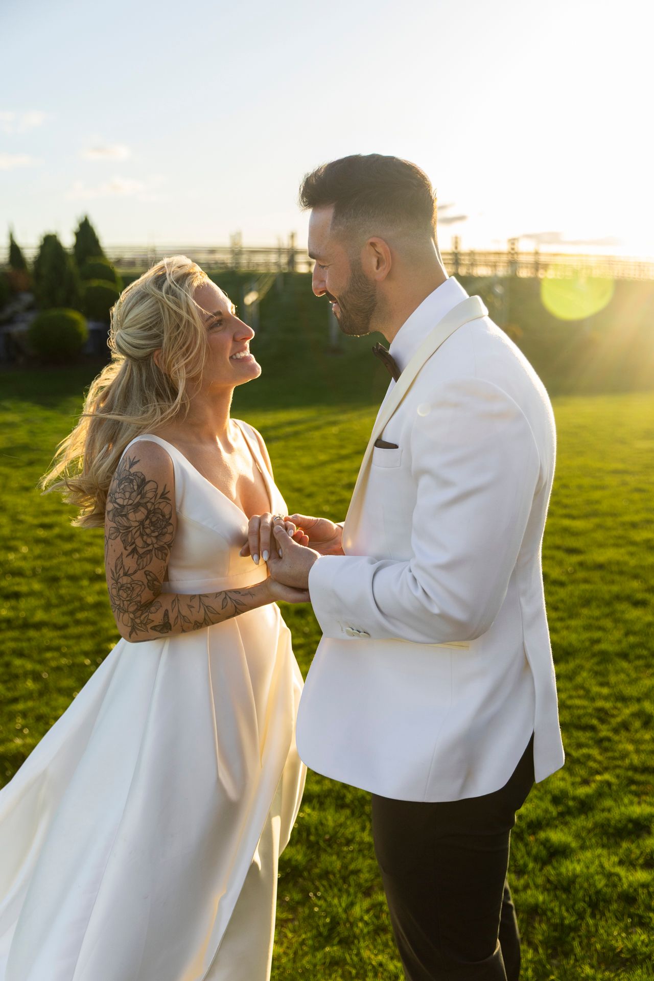Laughing bride and tattooed groom in white tuxedo hold hands on a sunlit green lawn at sunset.