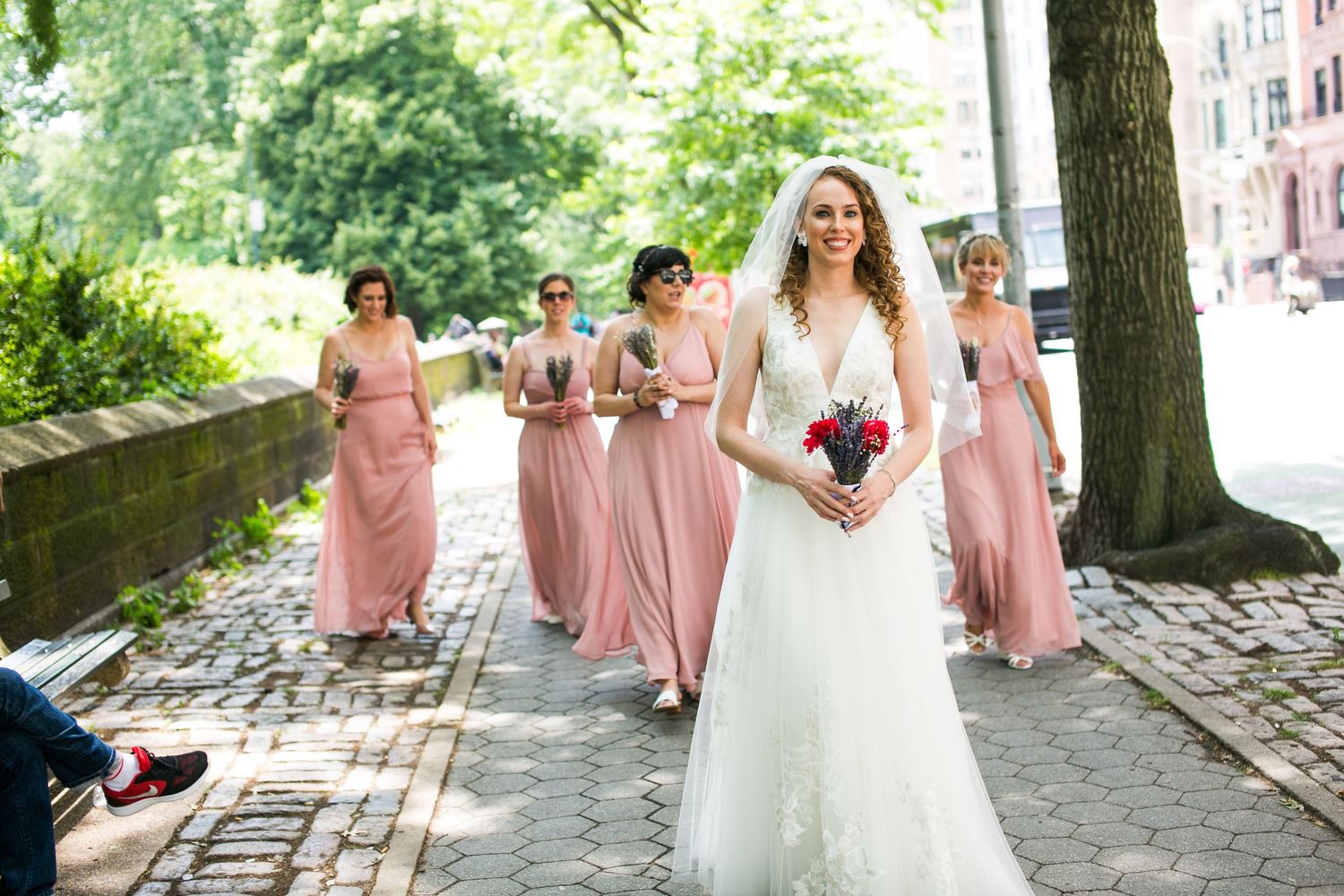 Bride in white gown leads bridesmaids in pink dresses along a cobblestone path surrounded by trees.
