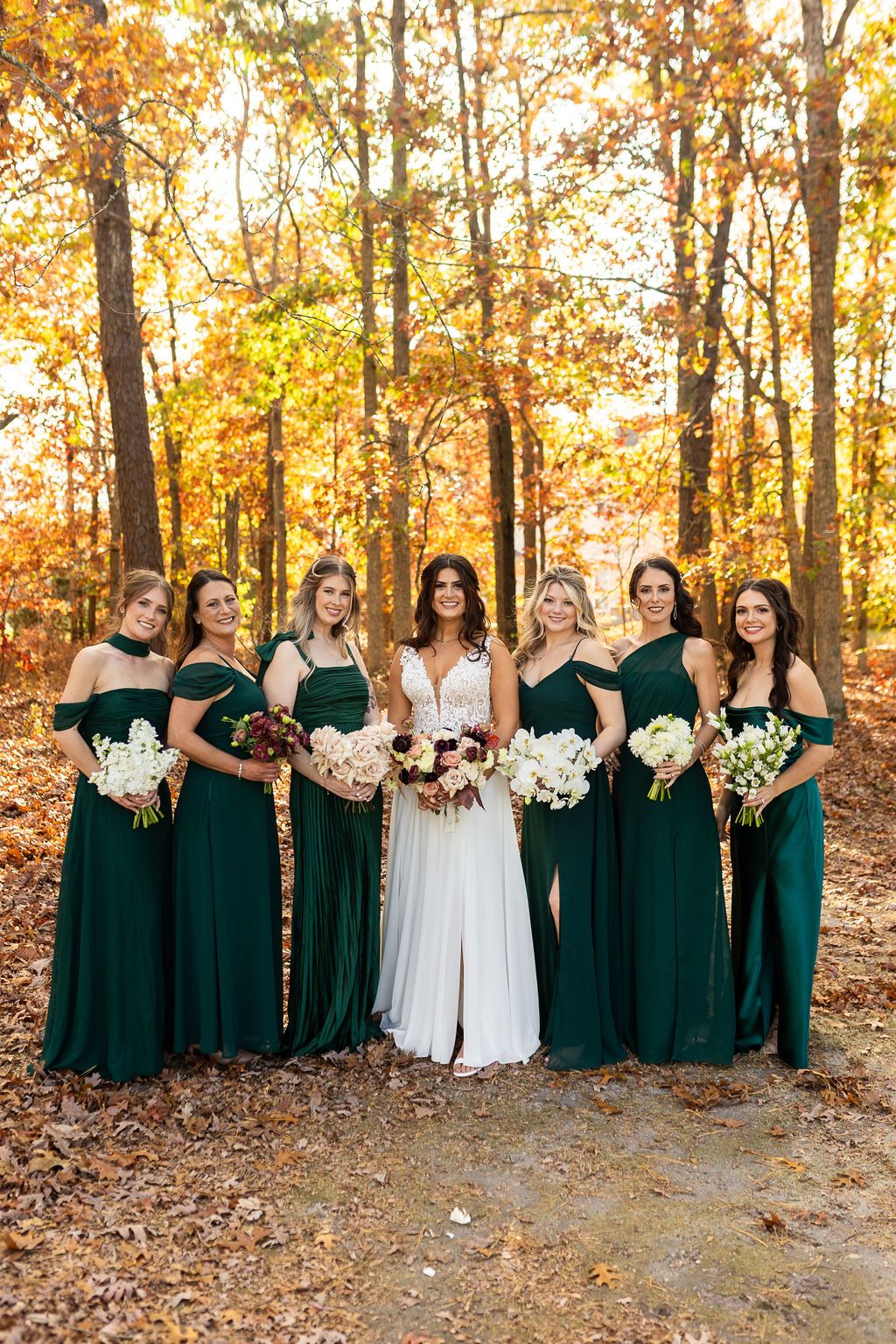 Bride in white gown posing with six bridesmaids in emerald green dresses holding bouquets in fall forest.
