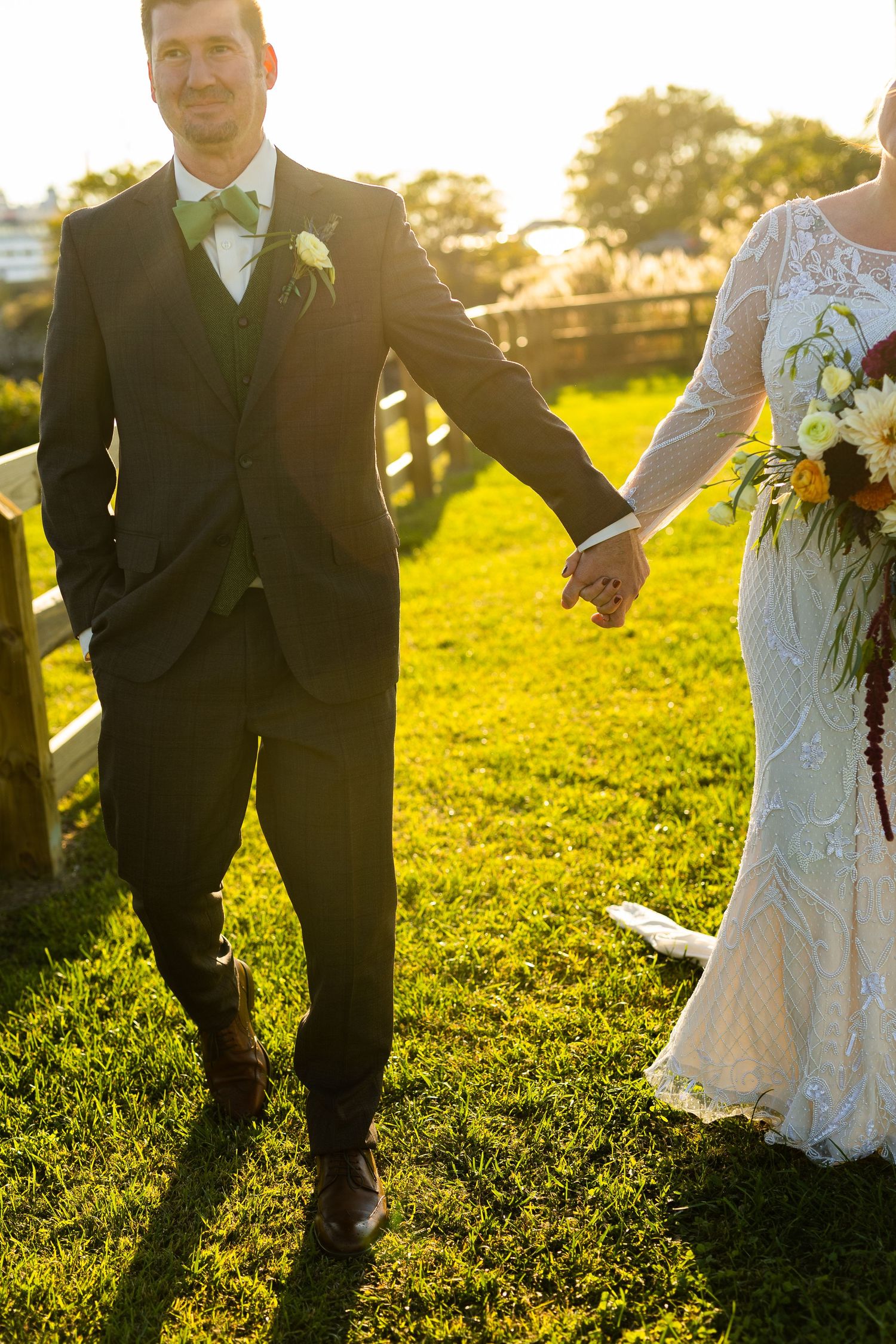 Bride and groom holding hands walking through sun-drenched green field at sunset.