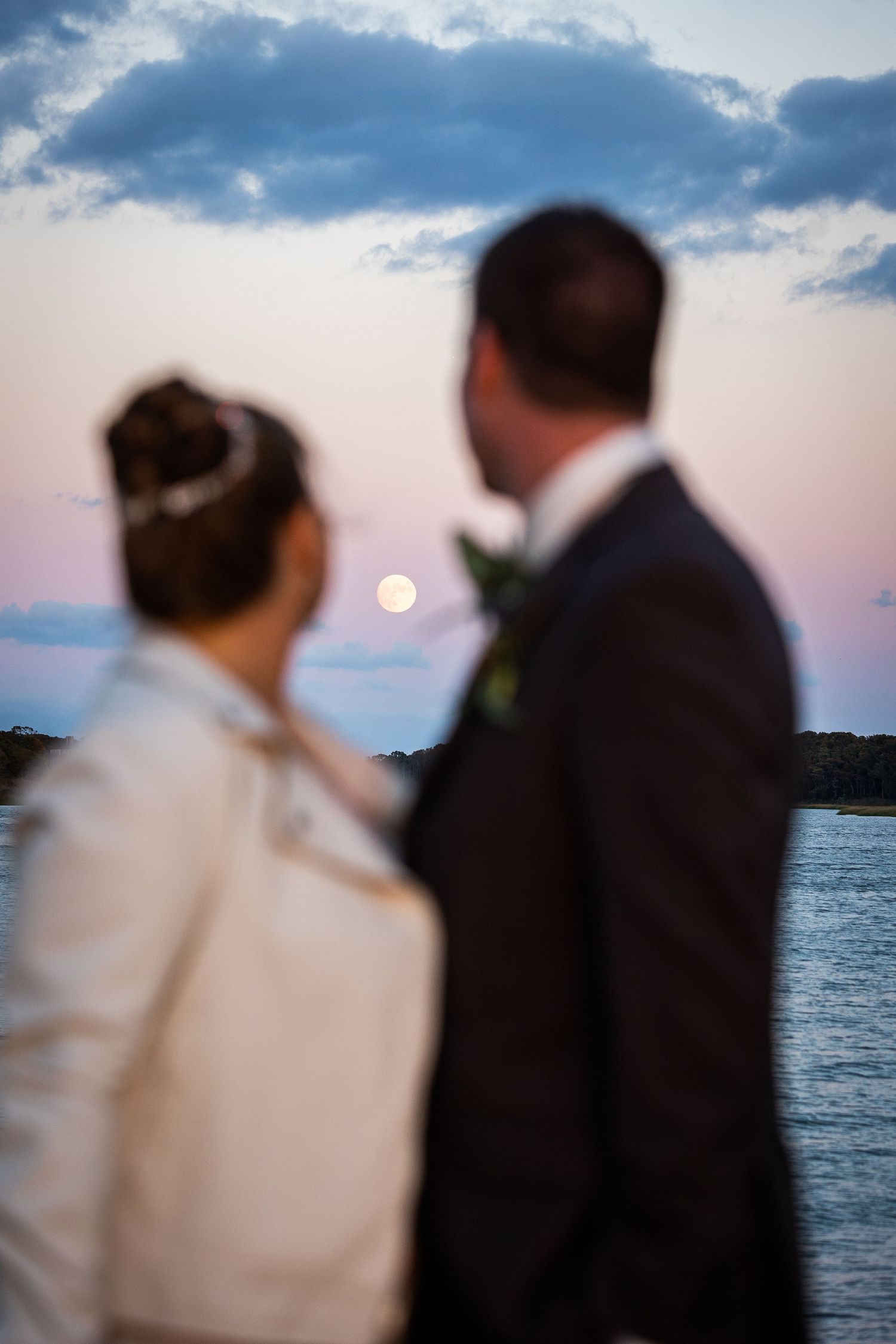 Bride and groom share a romantic moment with a full moon rising over the water at sunset.