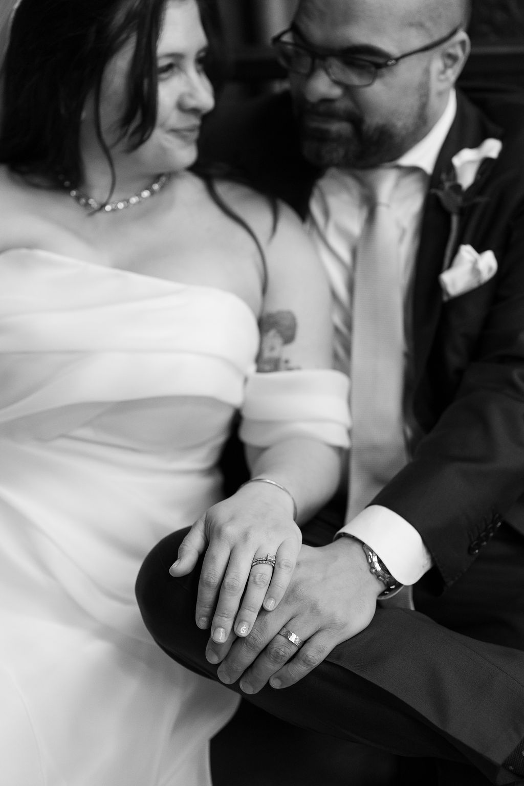 Close-up black and white photo of newlyweds displaying wedding rings on grooms knee at their wedding.
