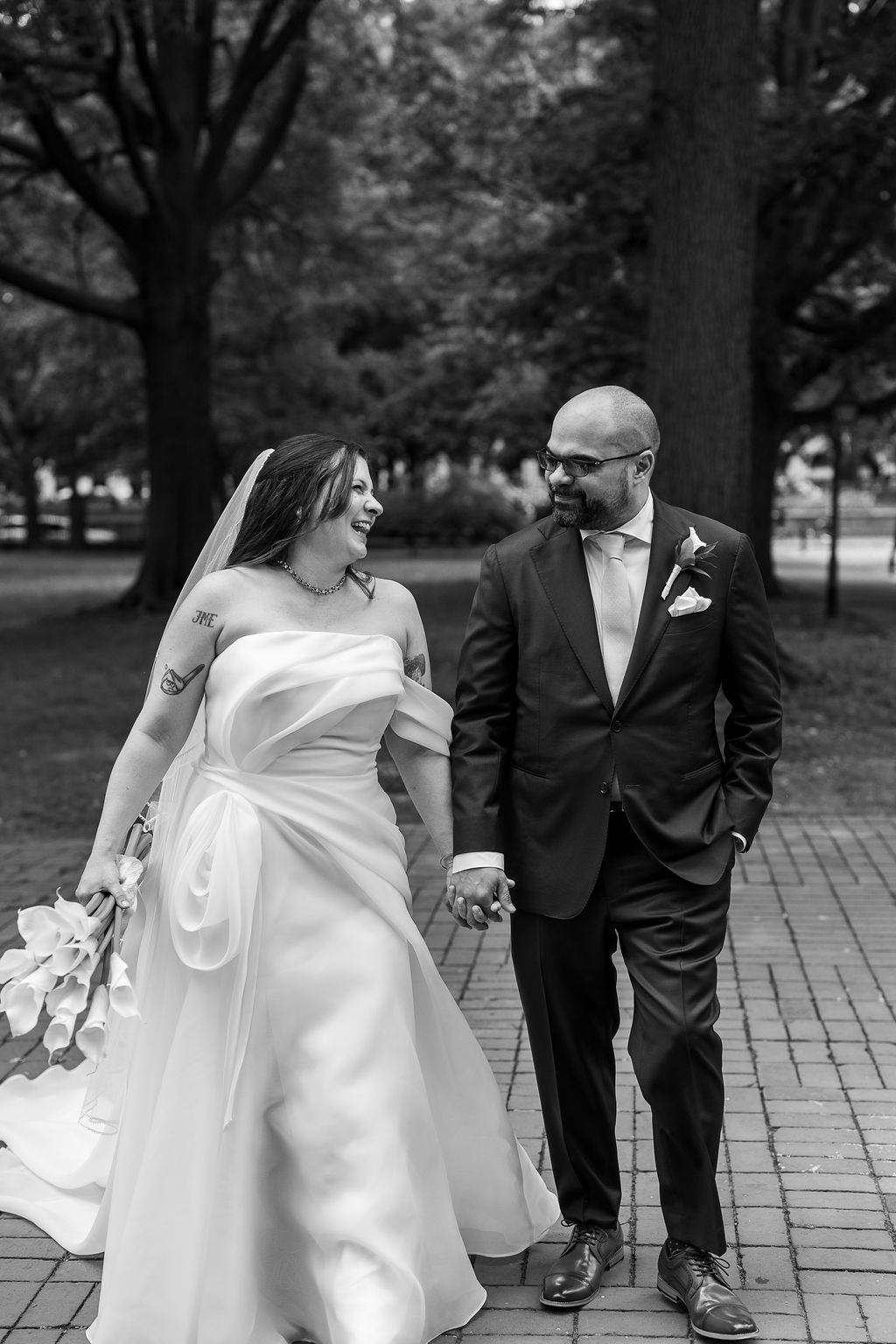 Black and white photo of laughing bride and smiling groom holding hands while walking in the park.