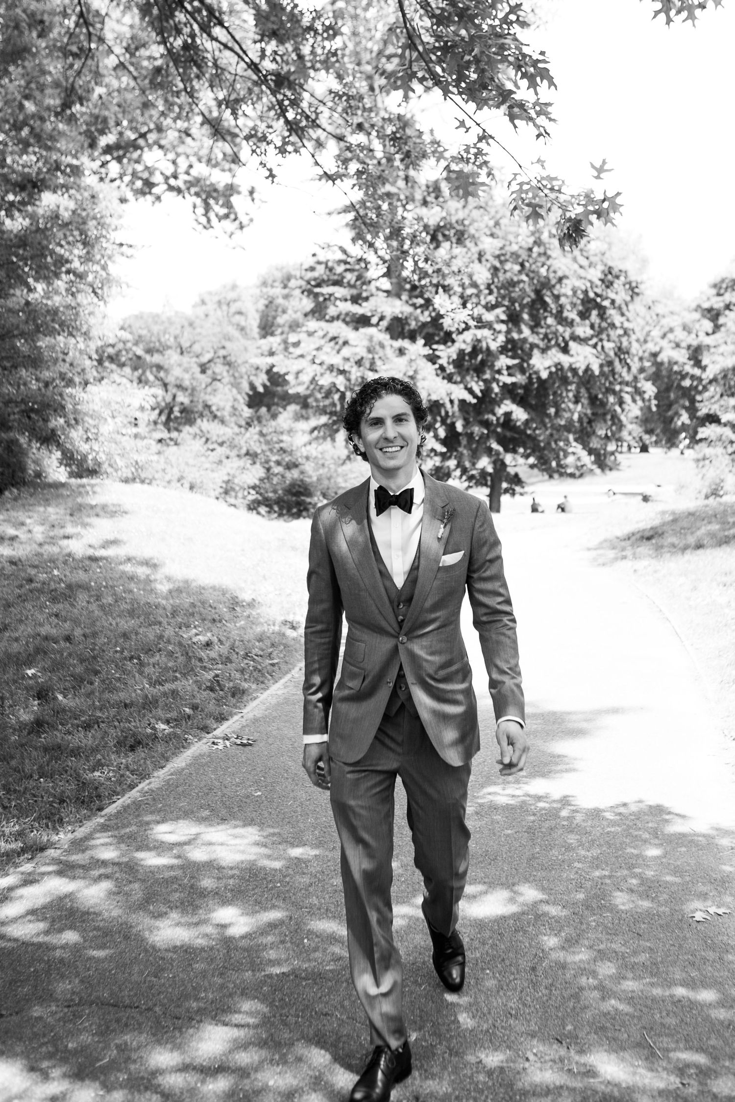 Groom in a gray suit and bow tie walking along a park path in black and white photo.