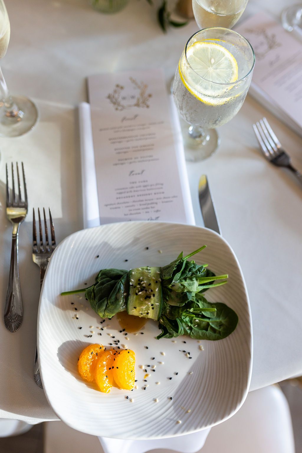 Elegant wedding reception first course plate with greens and orange segments beside lemon water and menu card.