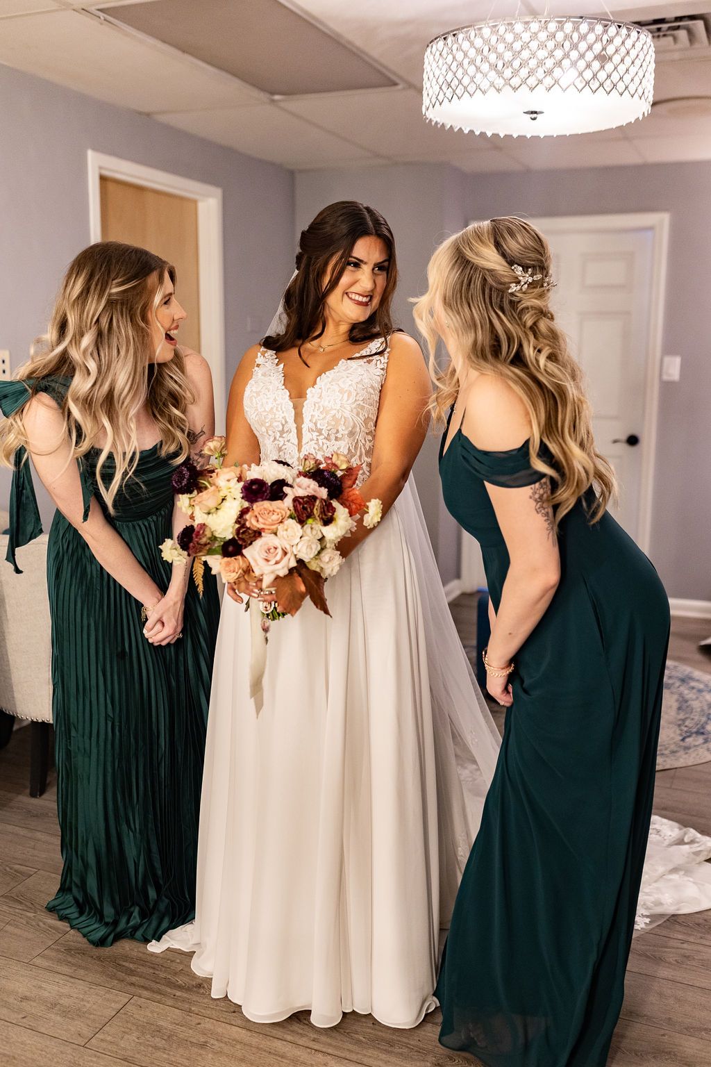 Bride in lace wedding gown holding a fall bouquet chatting with bridesmaids in emerald green dresses in a bridal suite.