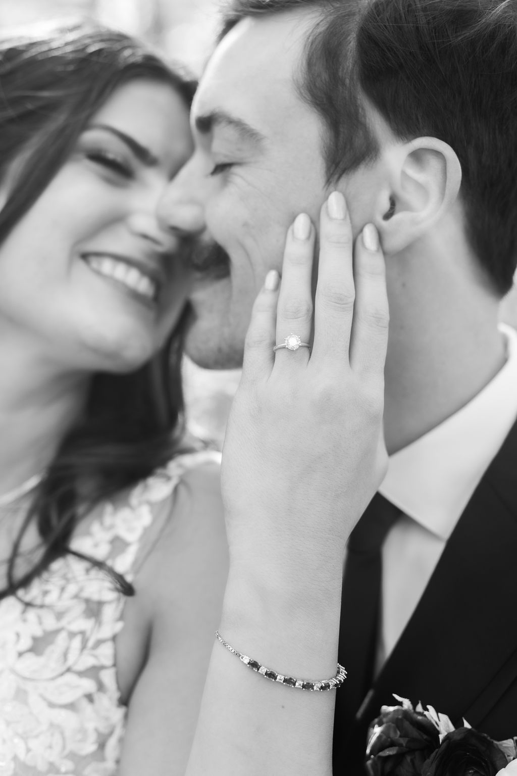 Black and white close-up of laughing bride showing wedding ring while touching grooms face tenderly.