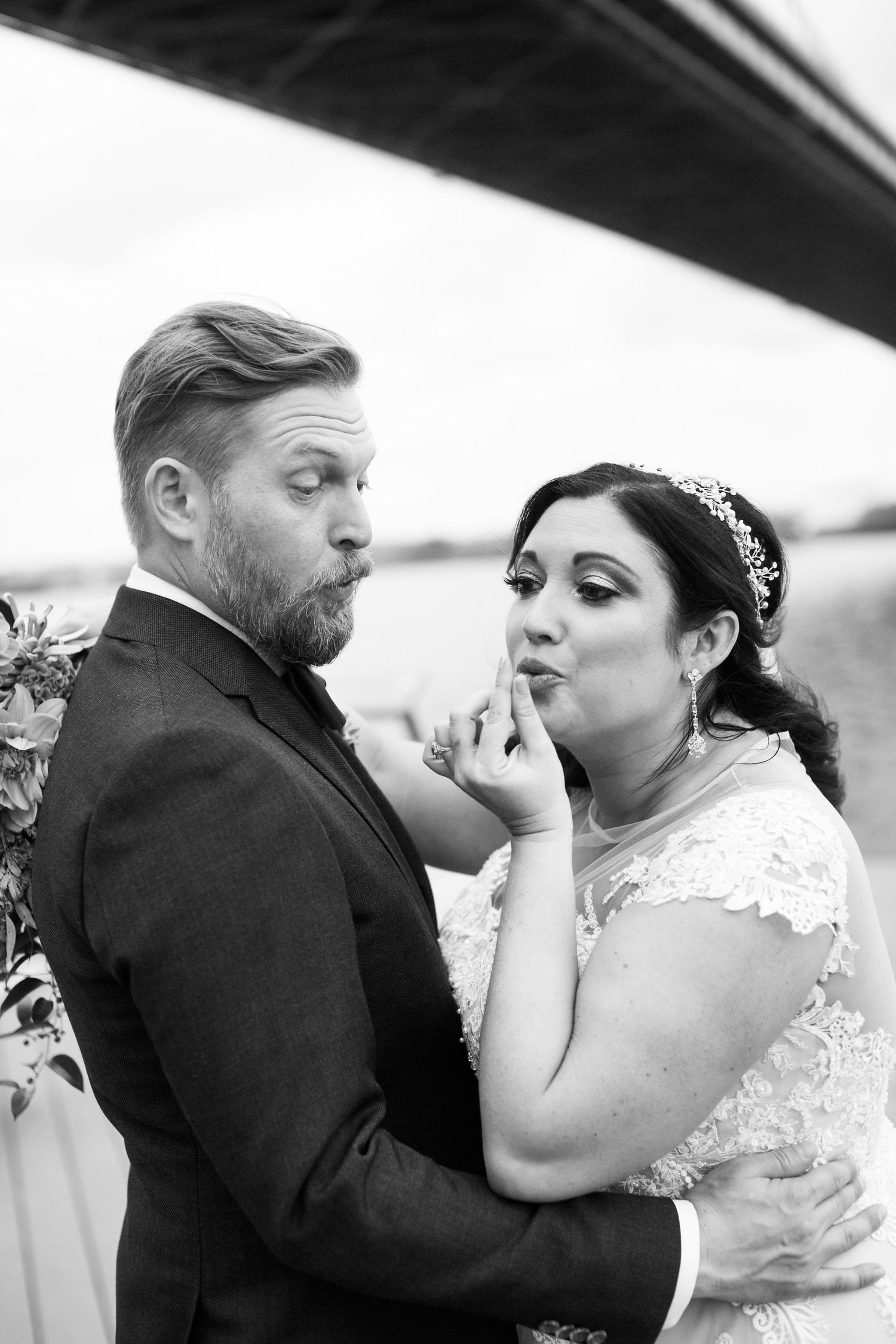 Black and white playful photo of bride touching grooms lips near a bridge on their wedding day.