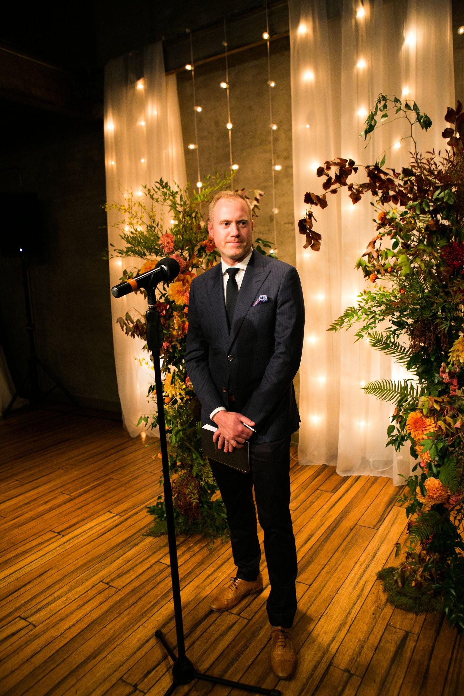 Officiant in navy suit stands at microphone before floral arch with fairy lights at wedding.