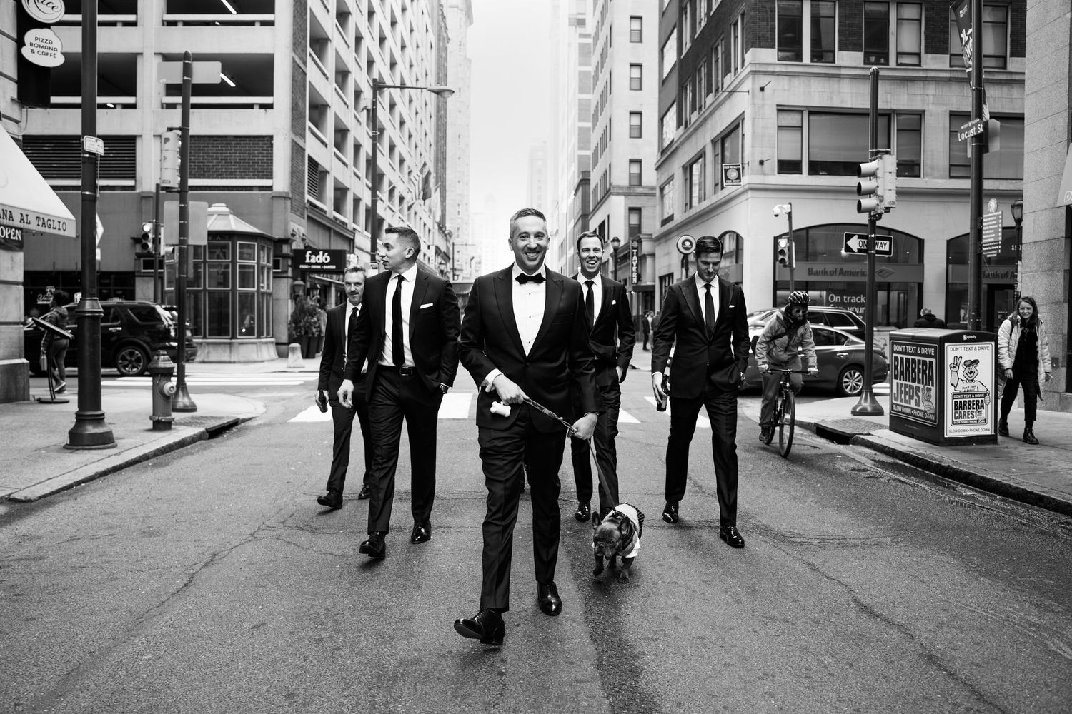 Groomsmen in black tuxedos walking down a city street with a French Bulldog in a black and white photo.