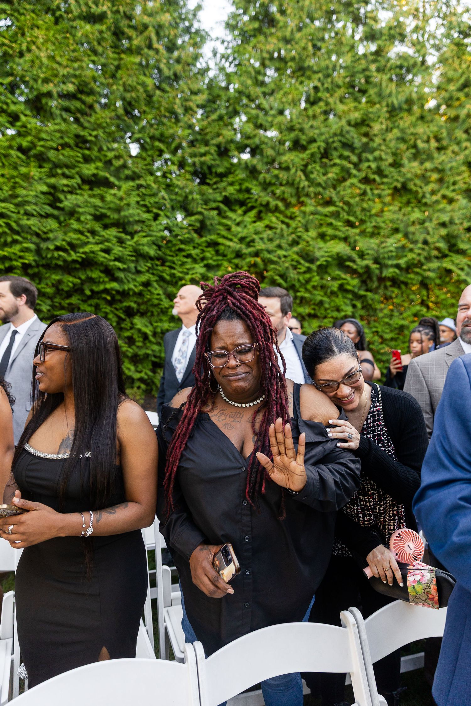 A woman with red locs in a black dress waves at an outdoor wedding ceremony surrounded by guests.