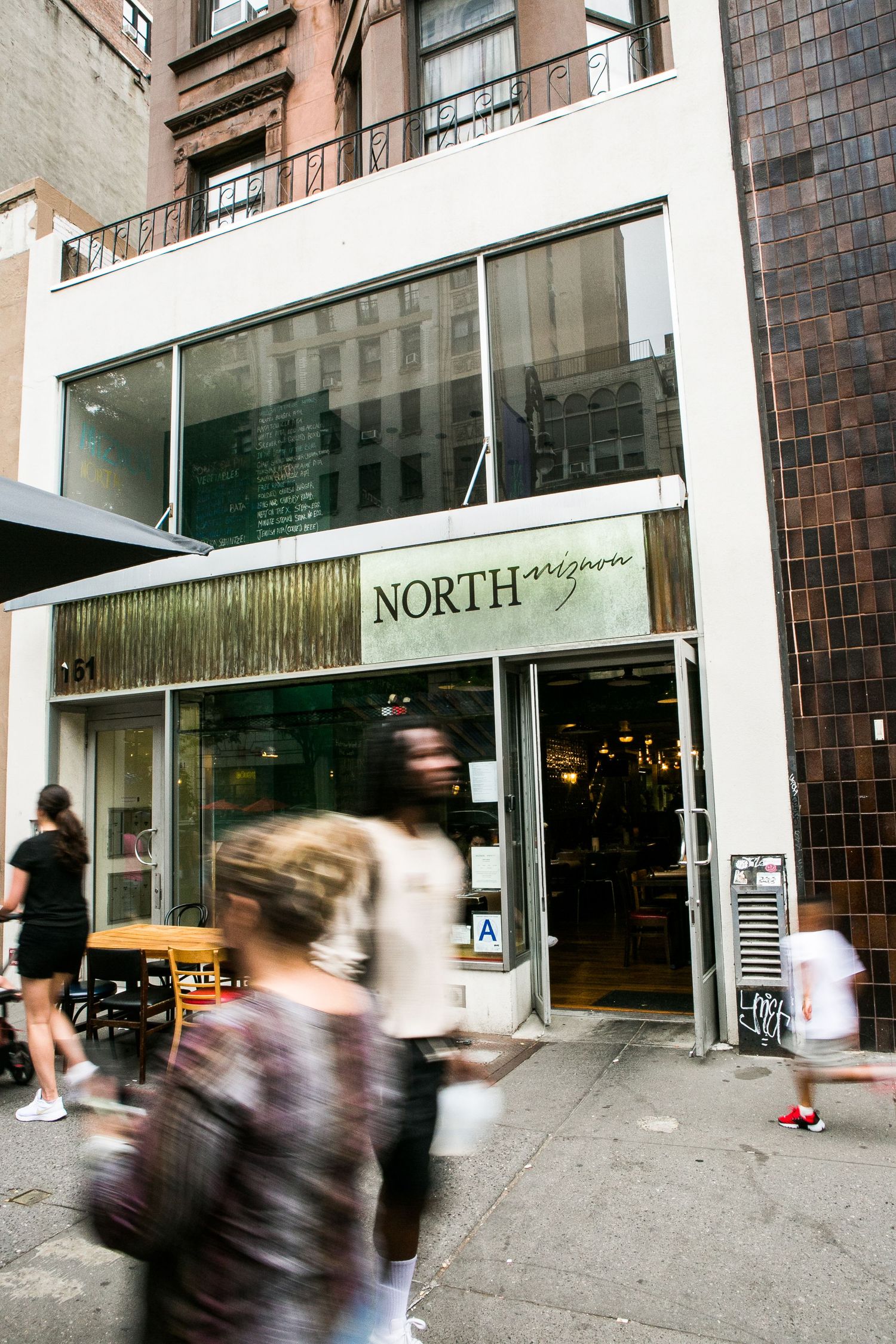 Pedestrians walk past North Mizzon restaurant storefront on a busy urban street corner in New York City.