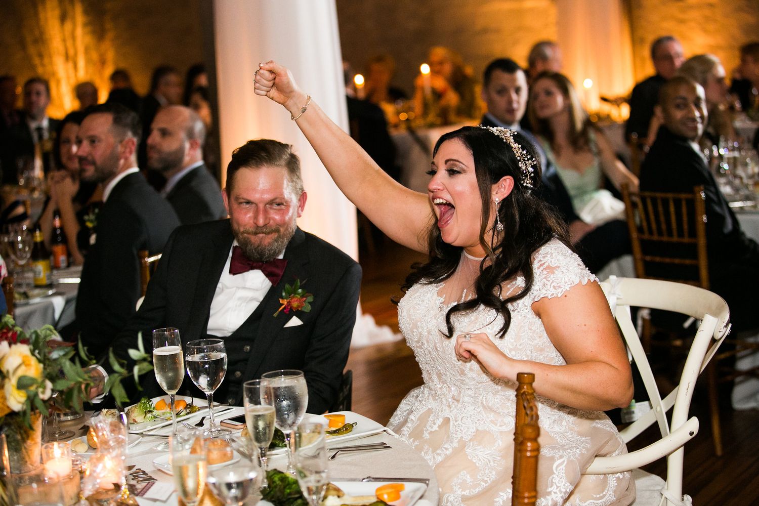 Bride cheering with raised fist at wedding reception dinner table.
