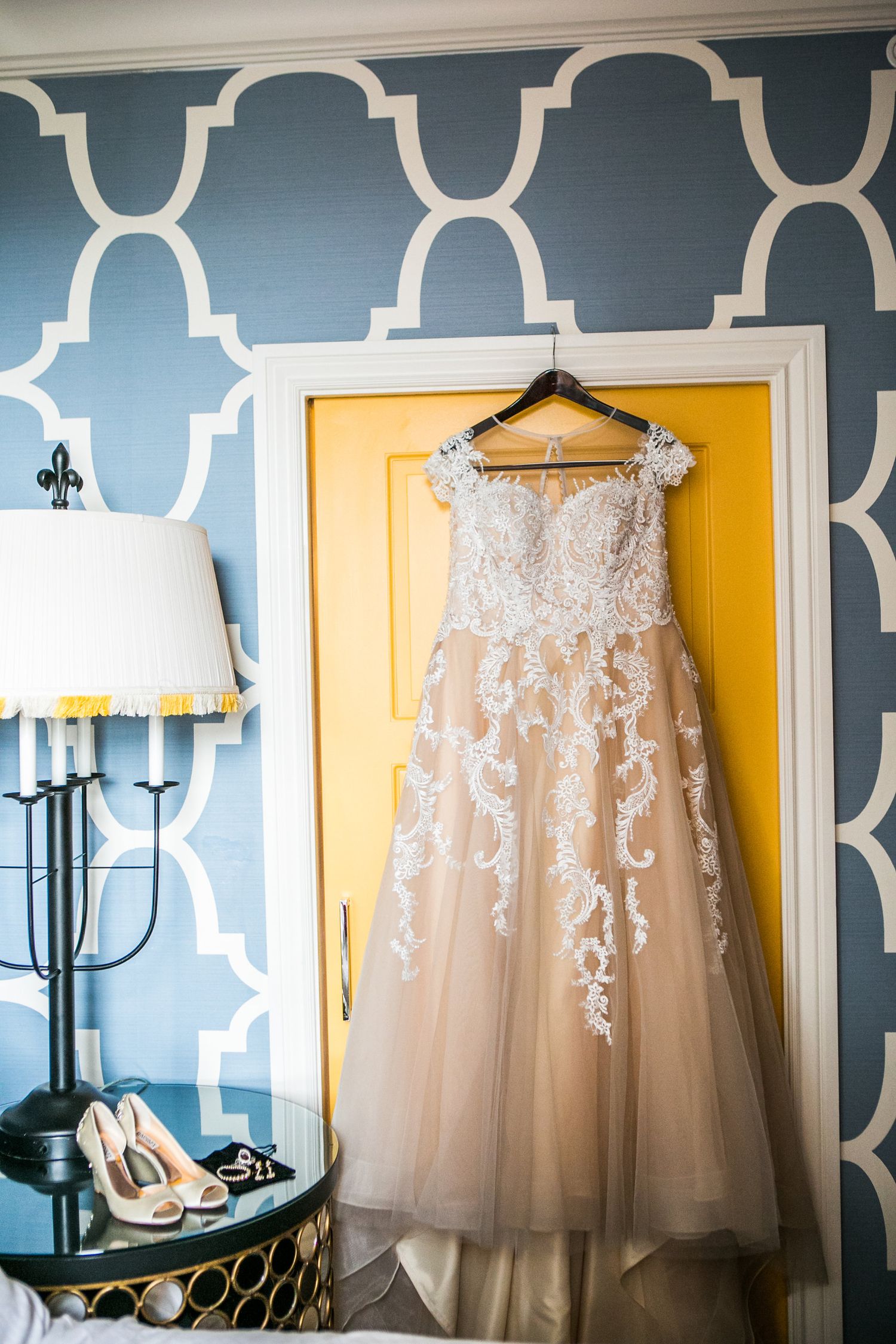 Elegant lace wedding gown hanging on yellow door frame in blue patterned bridal suite.
