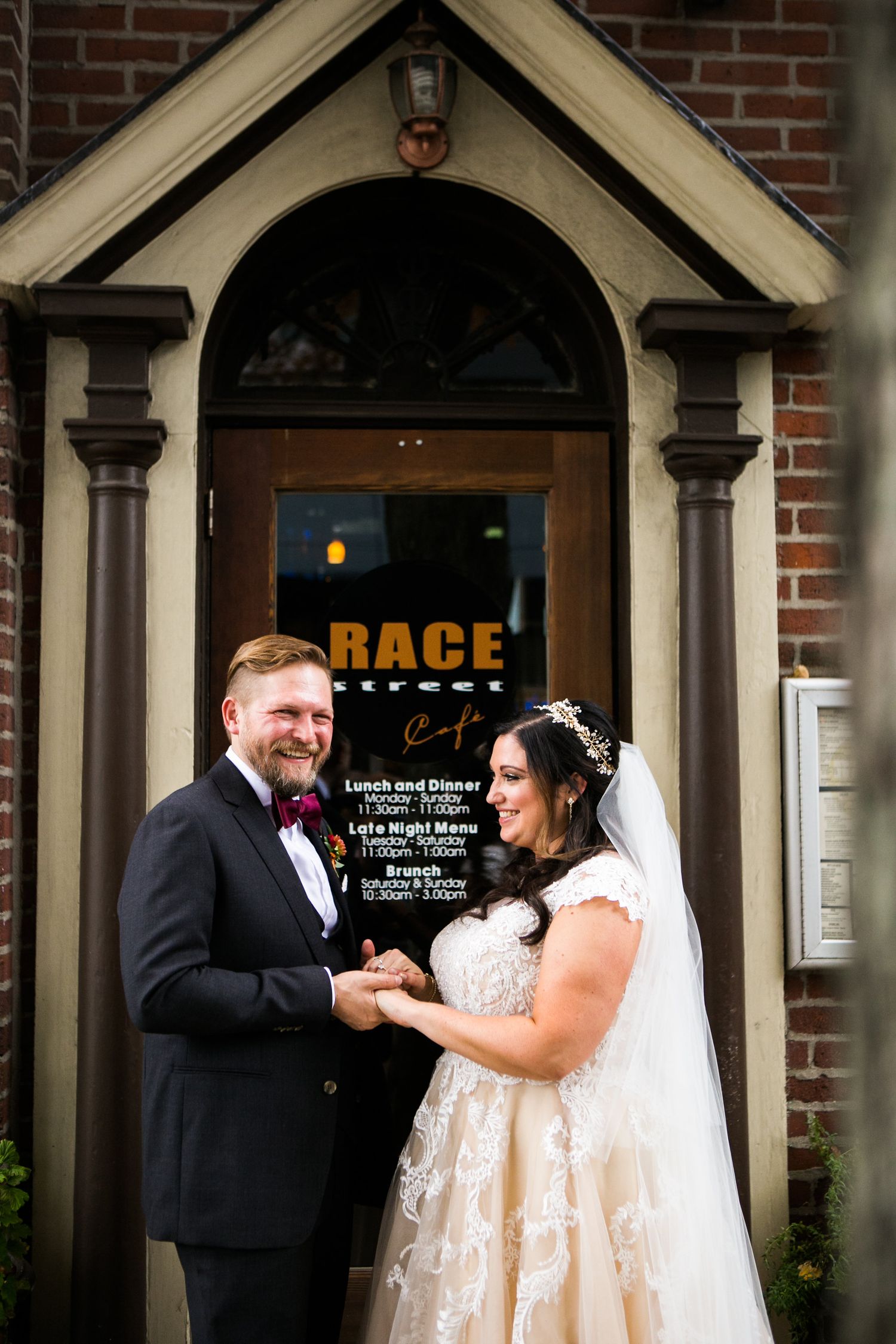 Smiling bride and groom hold hands outside Race Street Cafe, bride wearing lace ballgown and veil.
