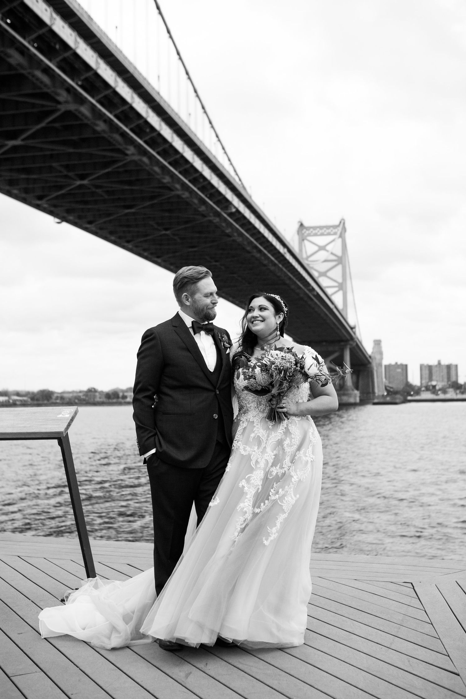 Black and white portrait of bride and groom standing on waterfront deck with Benjamin Franklin Bridge behind them.