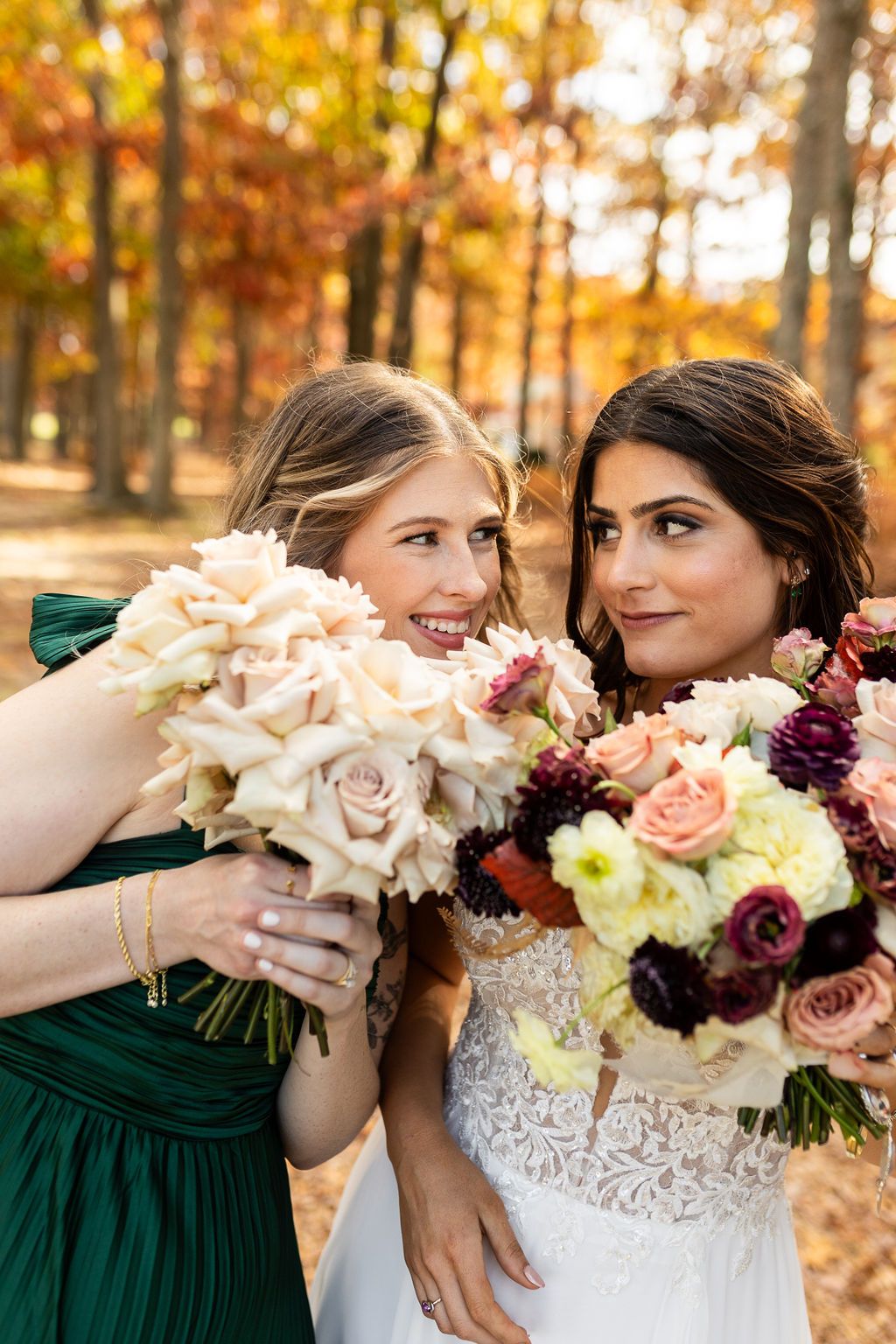 Bride in lace gown and bridesmaid in green dress holding floral bouquets among autumn foliage.