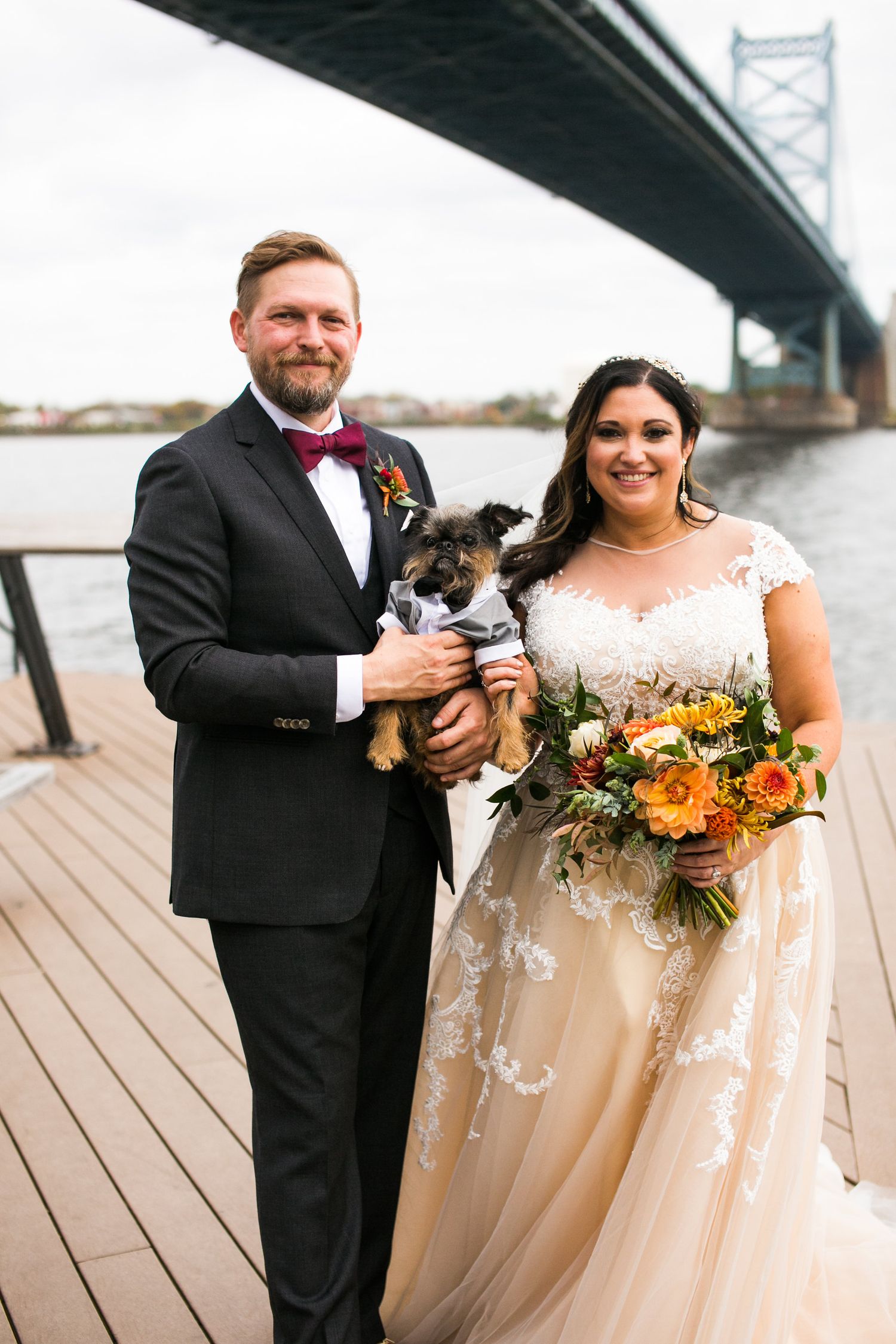 Bride and groom smile proudly holding their small dog in a suit on a waterfront dock by a bridge.