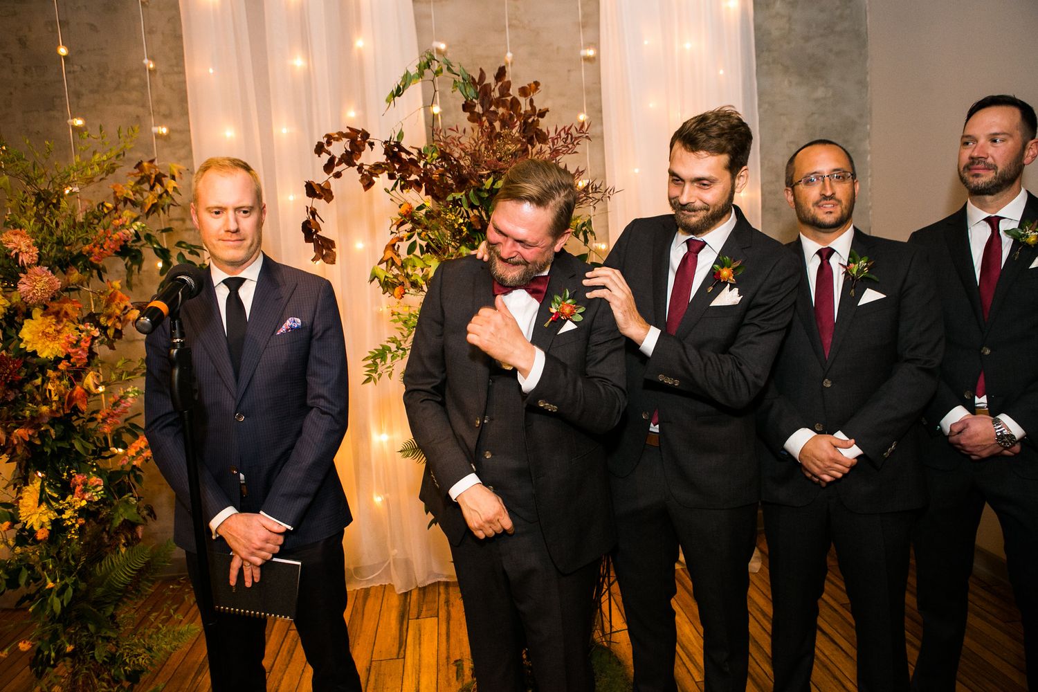 Emotional groom wipes tears surrounded by groomsmen in black suits at wedding altar.