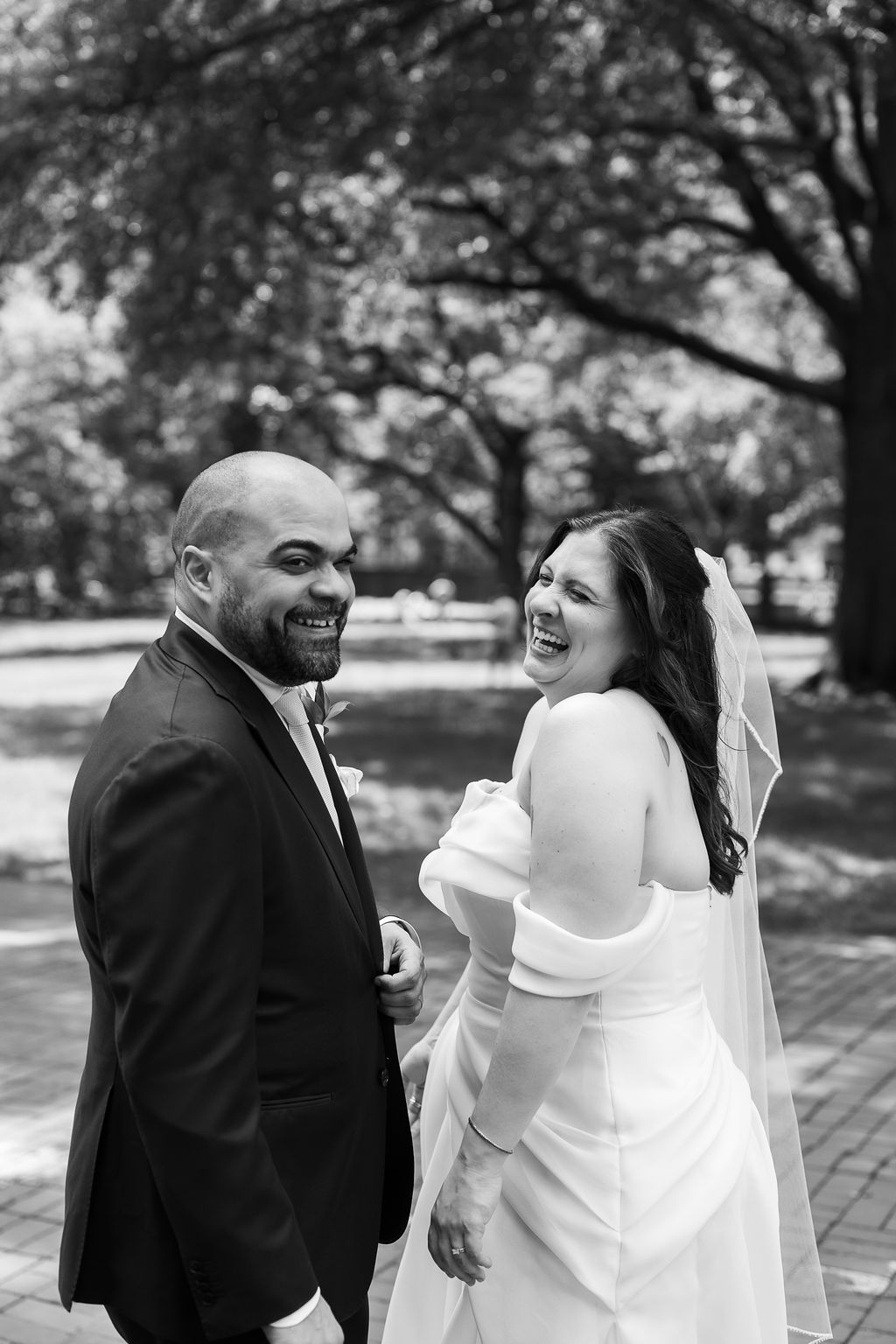 Bride and groom laugh joyfully together during outdoor wedding portraits in a sun-dappled park.