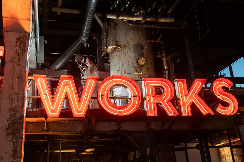 Glowing red neon WORKS sign mounted on rustic industrial building exterior with exposed pipes.