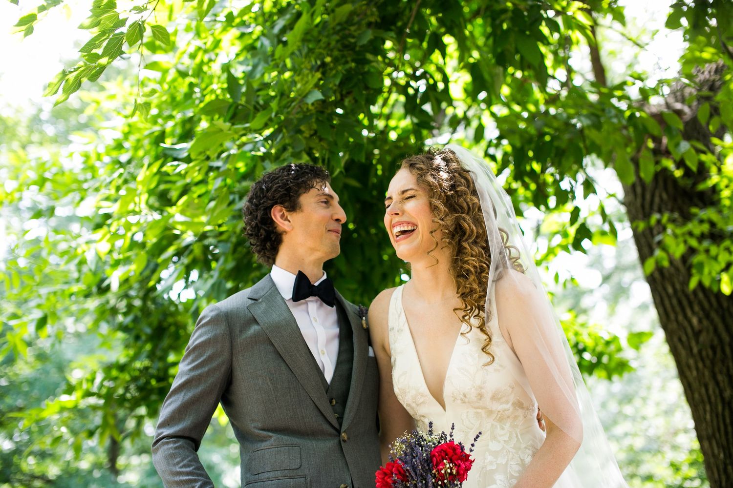 Smiling bride and groom share a joyful moment outdoors surrounded by lush green trees on their wedding day.