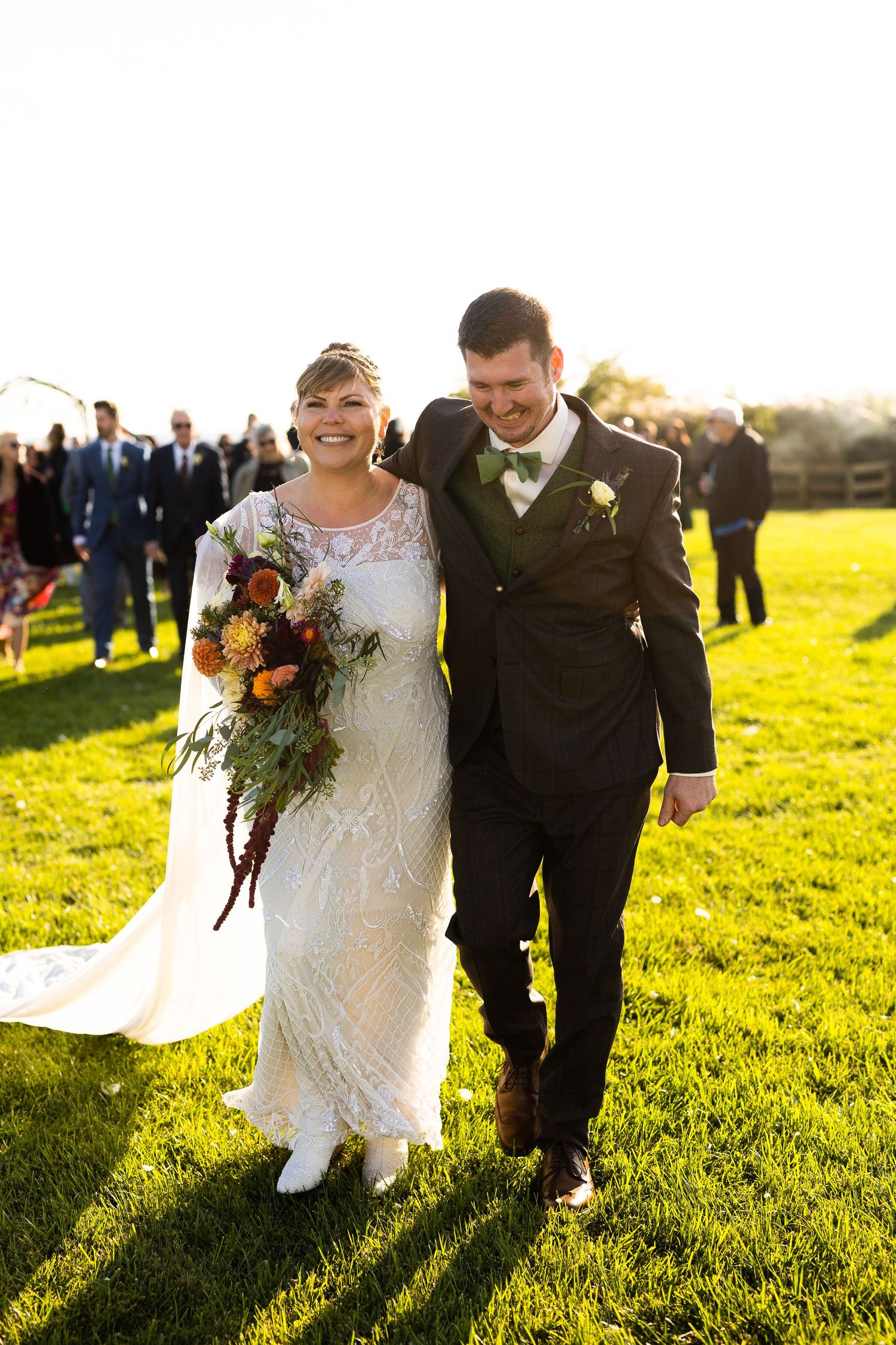 Joyful bride in lace gown and groom in dark suit walking hand-in-hand across green lawn after ceremony.