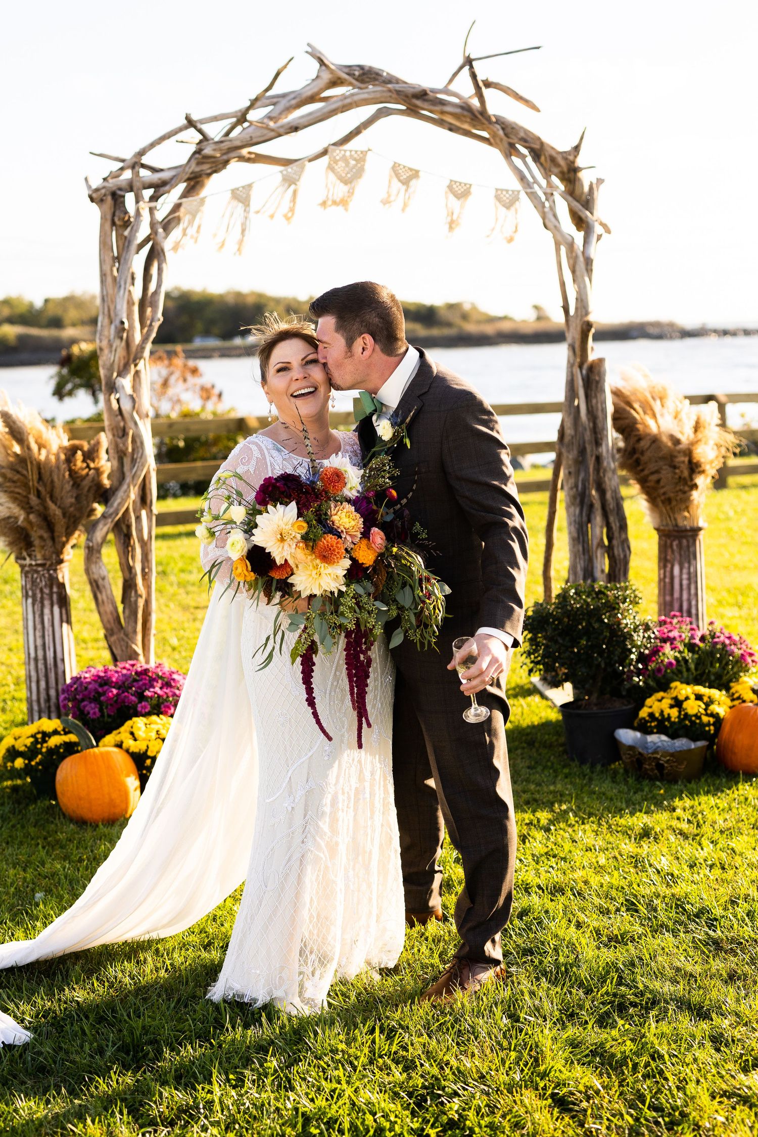 Bride and groom kiss under rustic driftwood arch decorated with pumpkins and fall flowers at waterfront wedding.