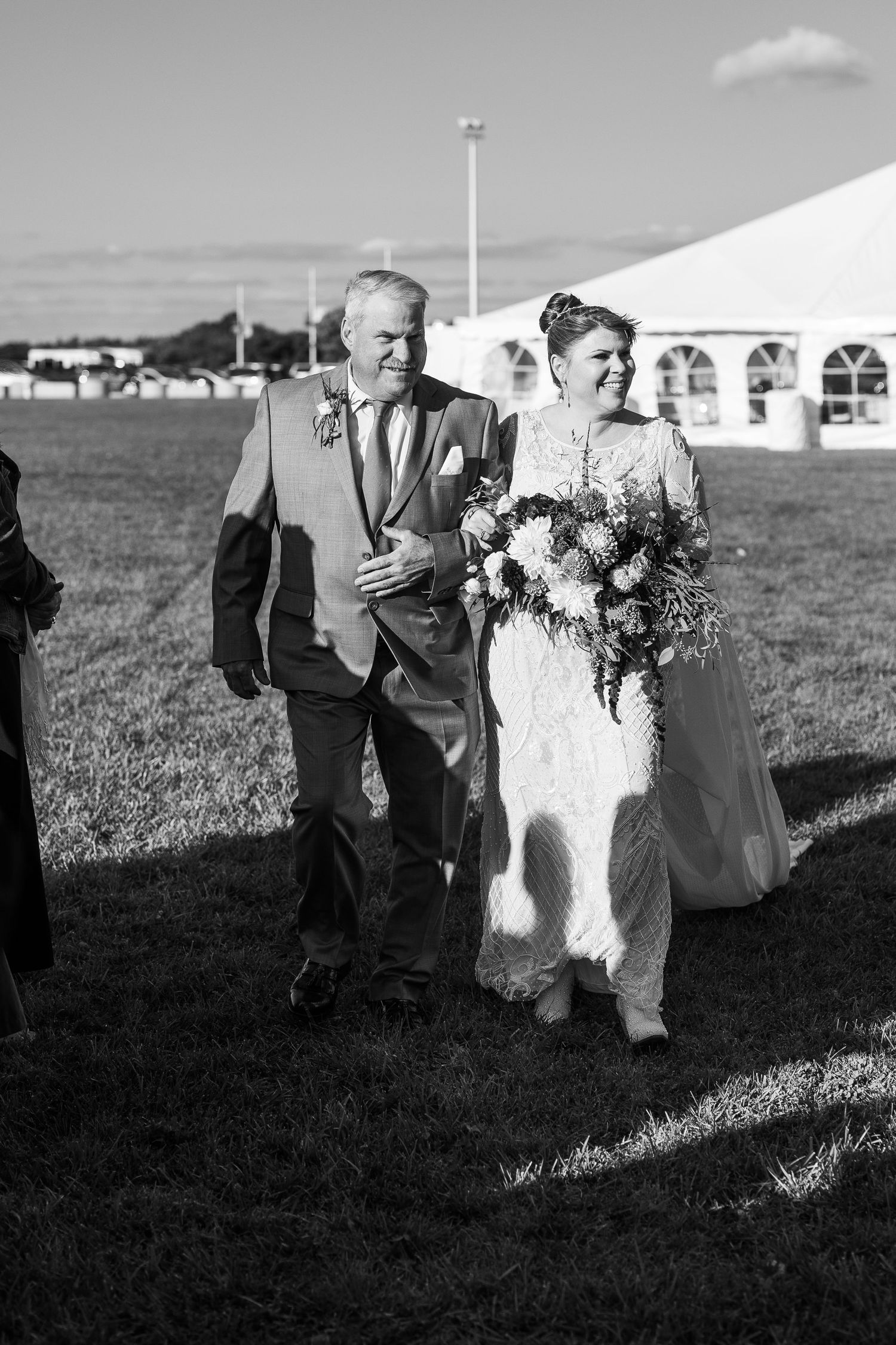Black and white photo of bride walking down the aisle with older man on grassy outdoor wedding venue.