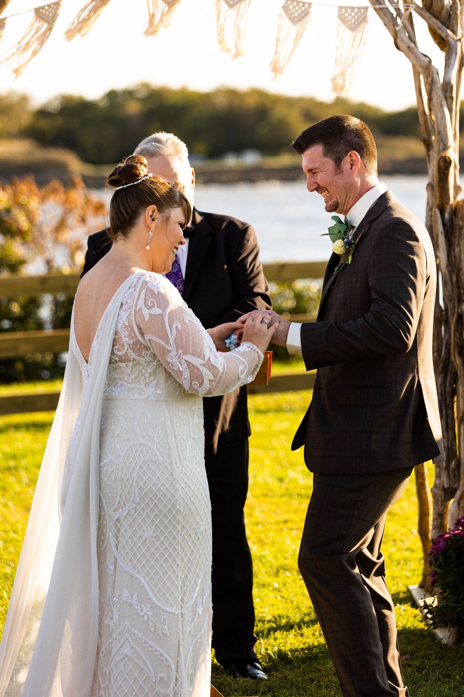 Bride placing wedding ring on groom's finger during outdoor ceremony by waterfront with macrame arch.