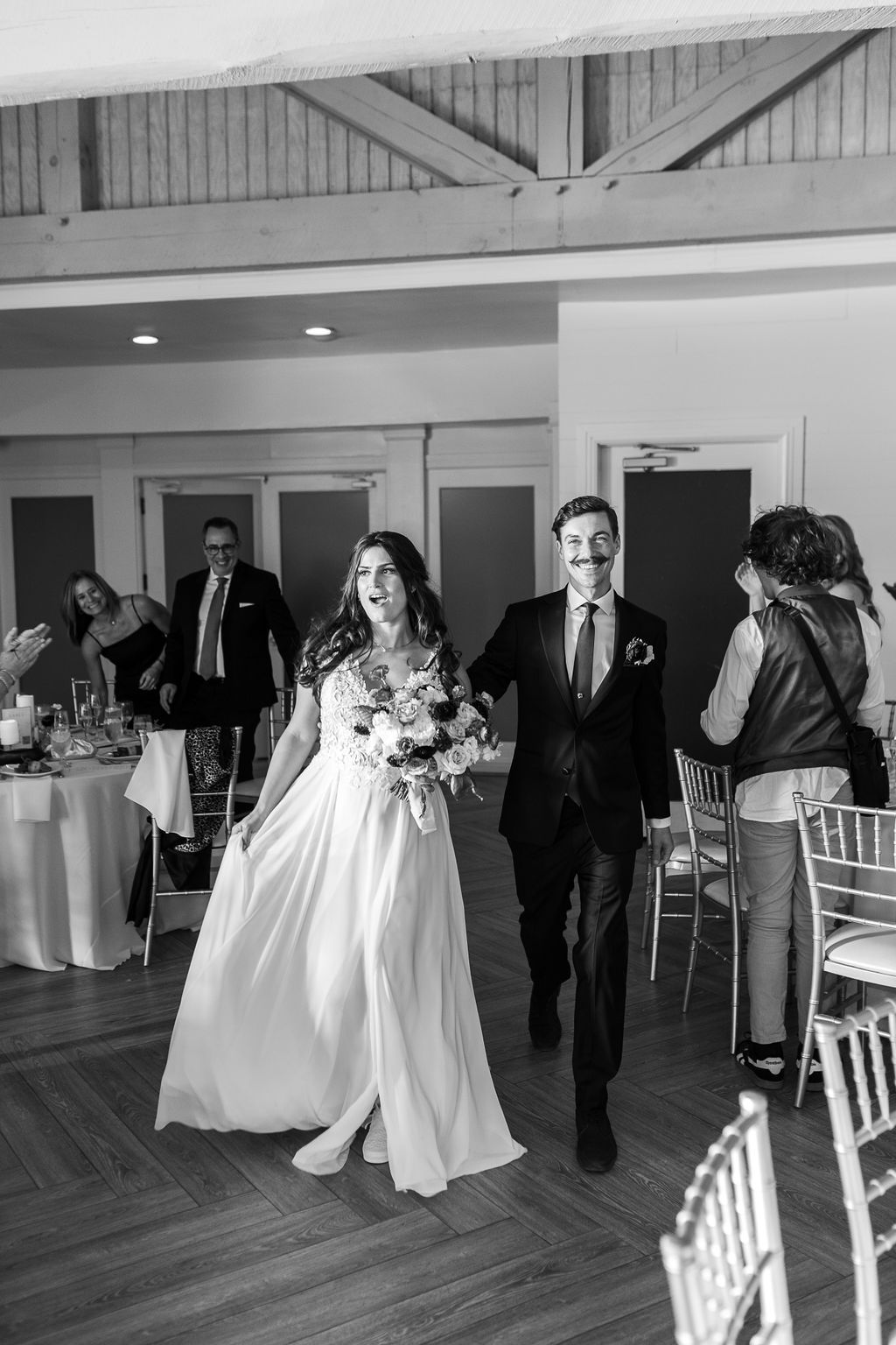 Bride and groom make joyful reception entrance in black and white photo, bride holding bouquet in flowing white gown.