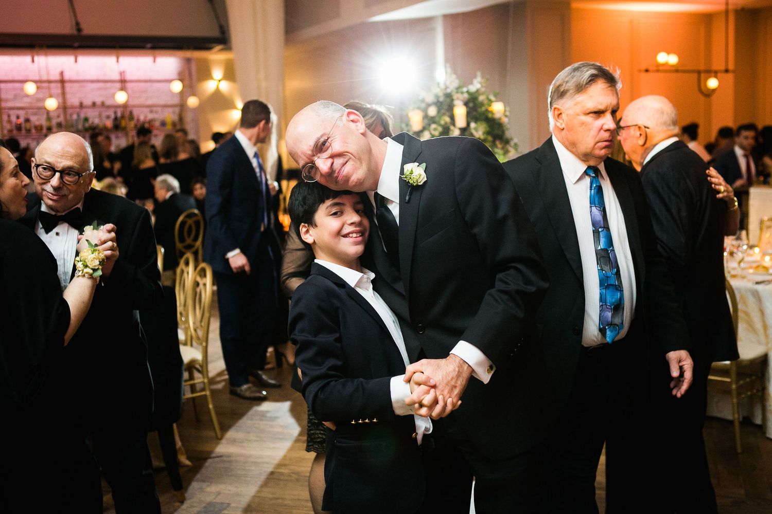 Older man in tuxedo dancing and posing with a young boy in a suit at a wedding reception.