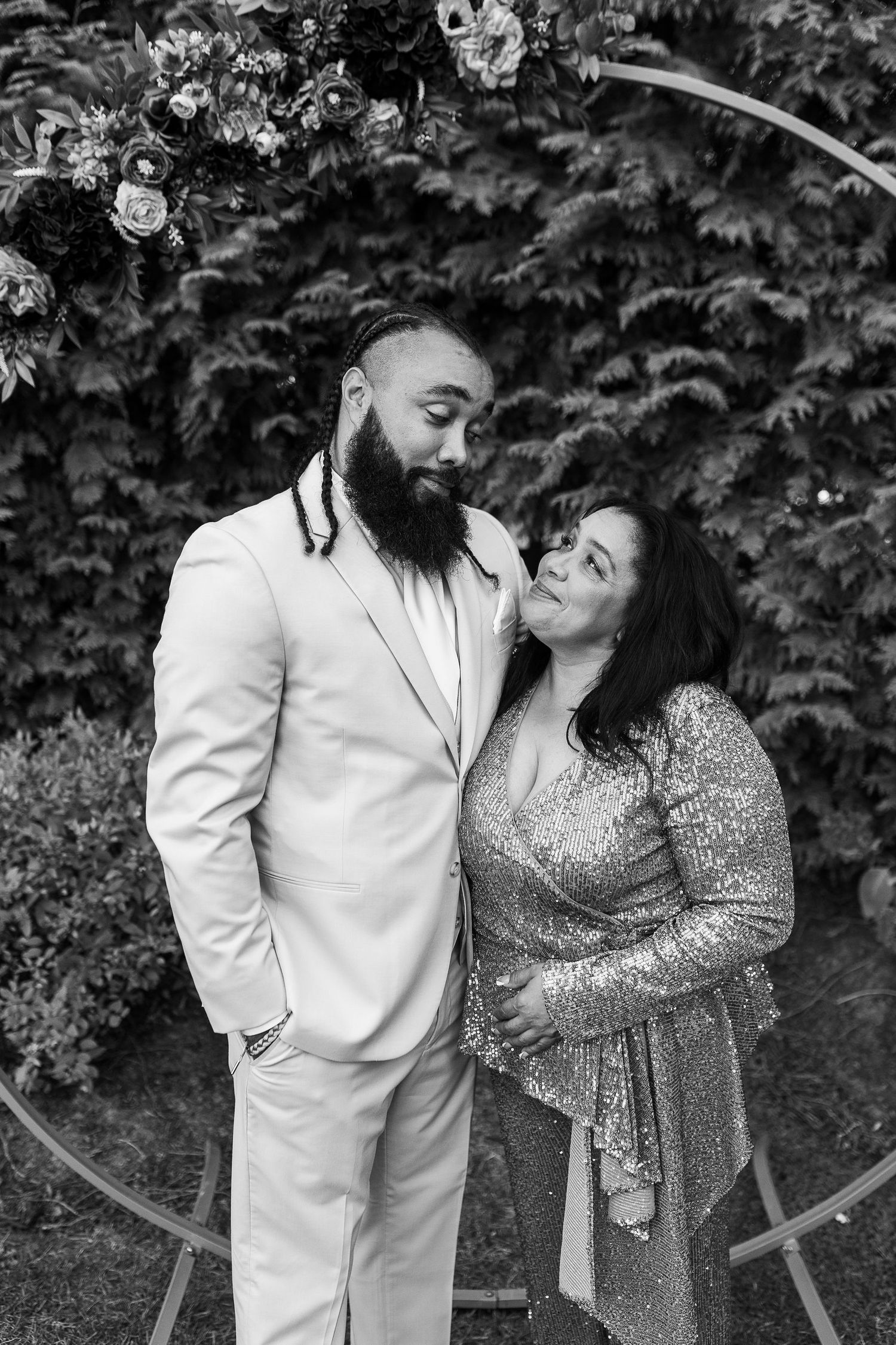 Black and white portrait of a bearded groom in a white suit with a woman in a sequined dress at a floral arch.