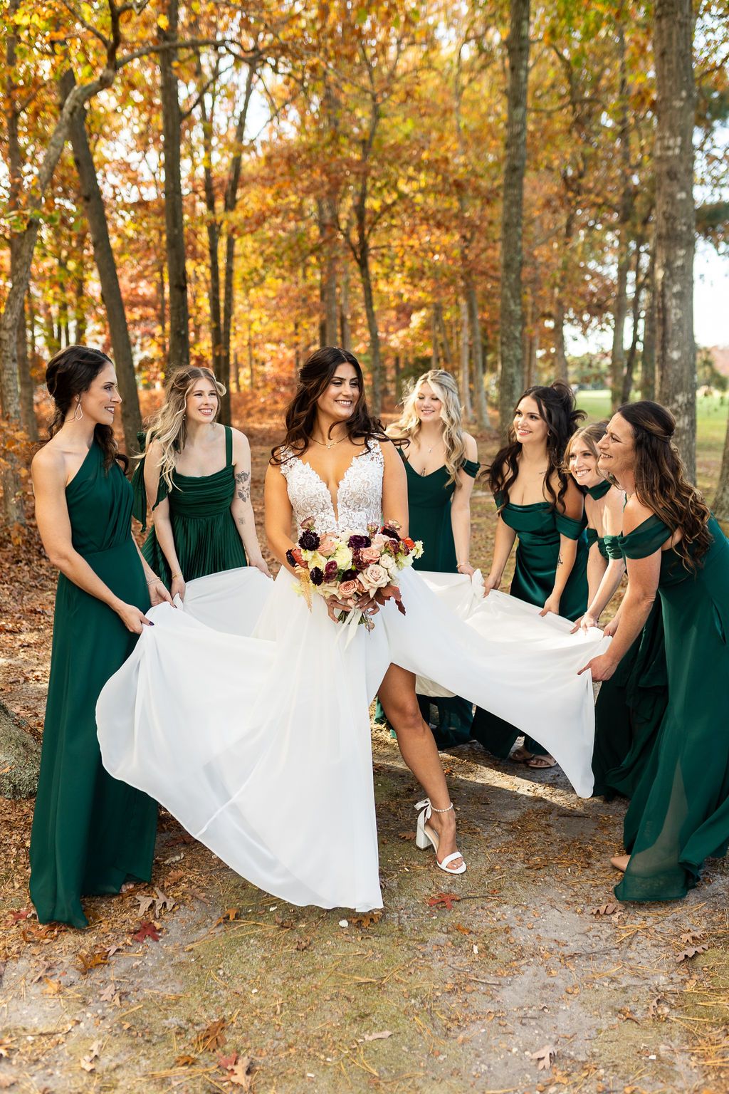 Bride holding bouquet surrounded by bridesmaids in emerald green gowns lifting her skirt among autumn trees.