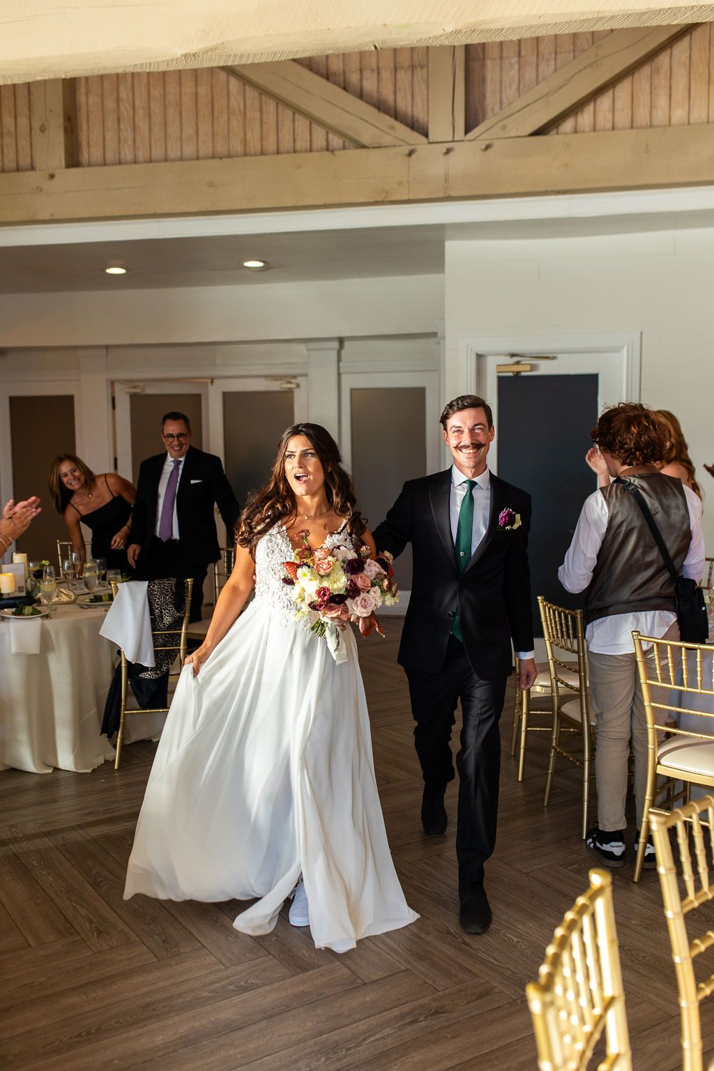 Smiling bride and groom enter wedding reception, bride carrying colorful bouquet in elegant white gown.