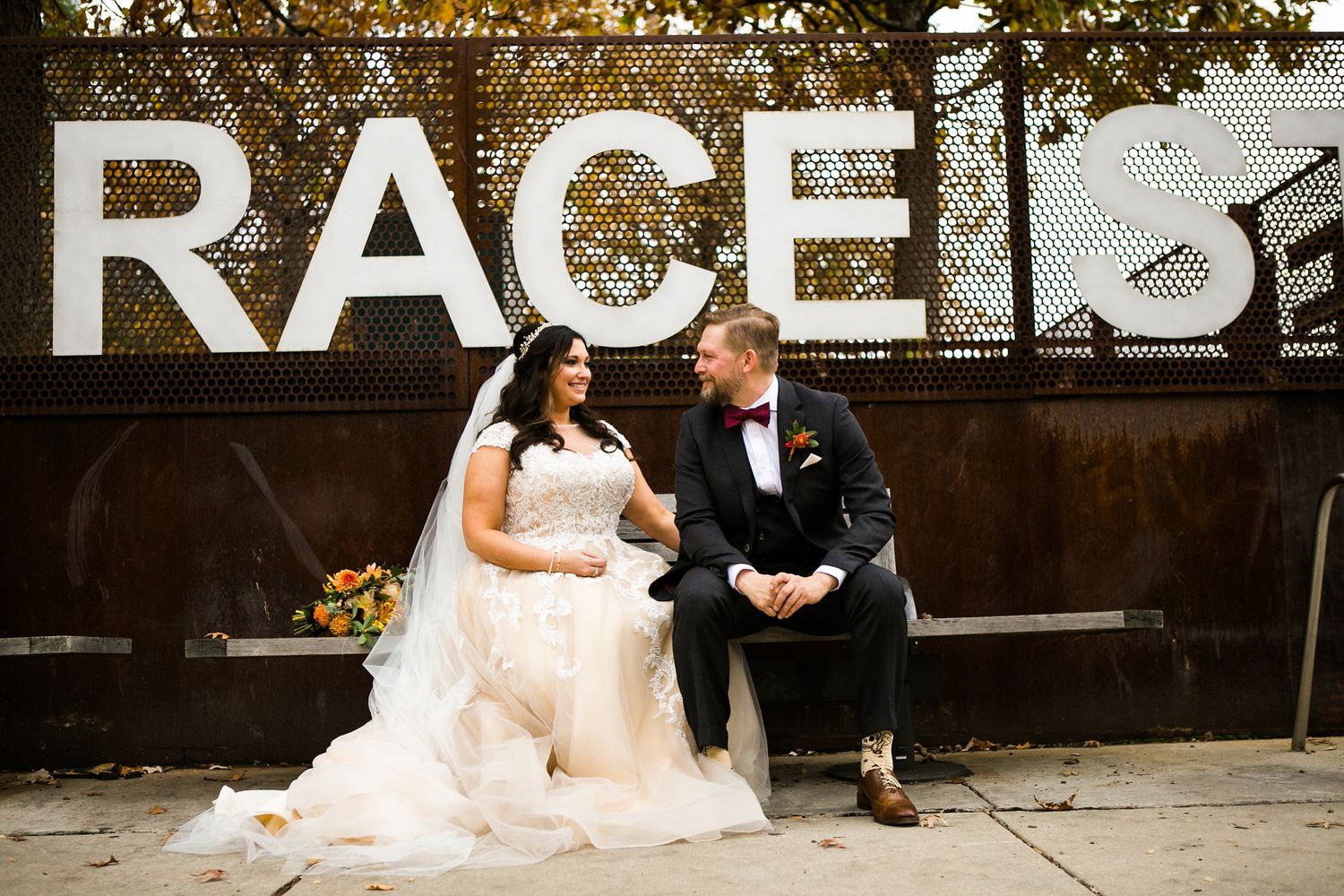 Bride in lace gown and groom in tuxedo share a romantic moment at Race Street sign in color photo.