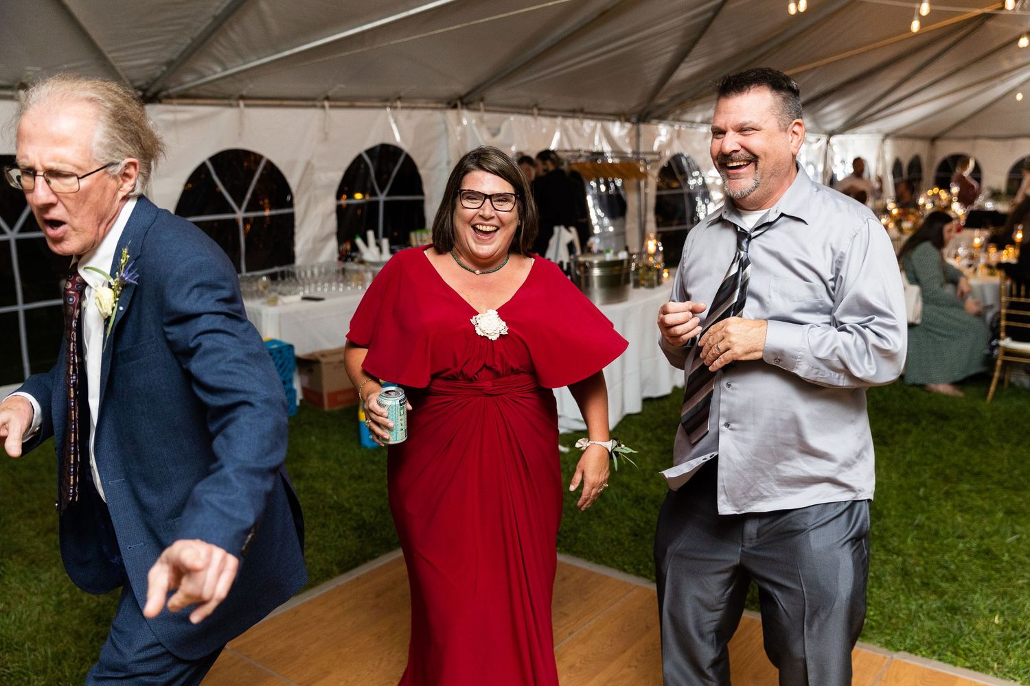 Laughing wedding guests in red and grey walk together on dance floor at tented reception.